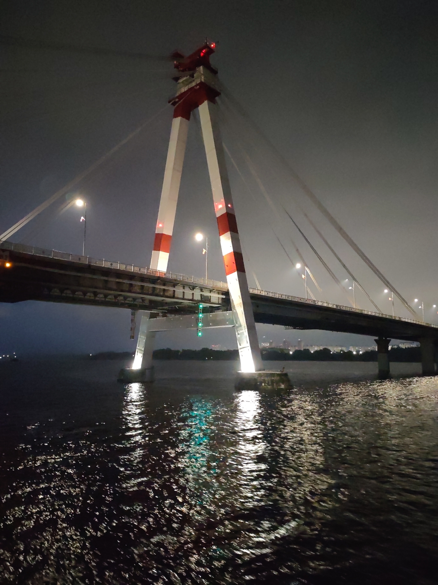 A nighttime view of the October Bridge in Cherepovets, Russia, dominates the scene. The bridge's architecture features tall, white, angled support towers with red accents at the top and middle sections. Cables extend from the top of the towers to the bridge deck. A section of the bridge, with streetlights visible along its length, spans over dark water. The water reflects light from the bridge supports and the green lights from beneath the bridge, creating a rippled effect. The sky is dark and overcast. No people are visible.