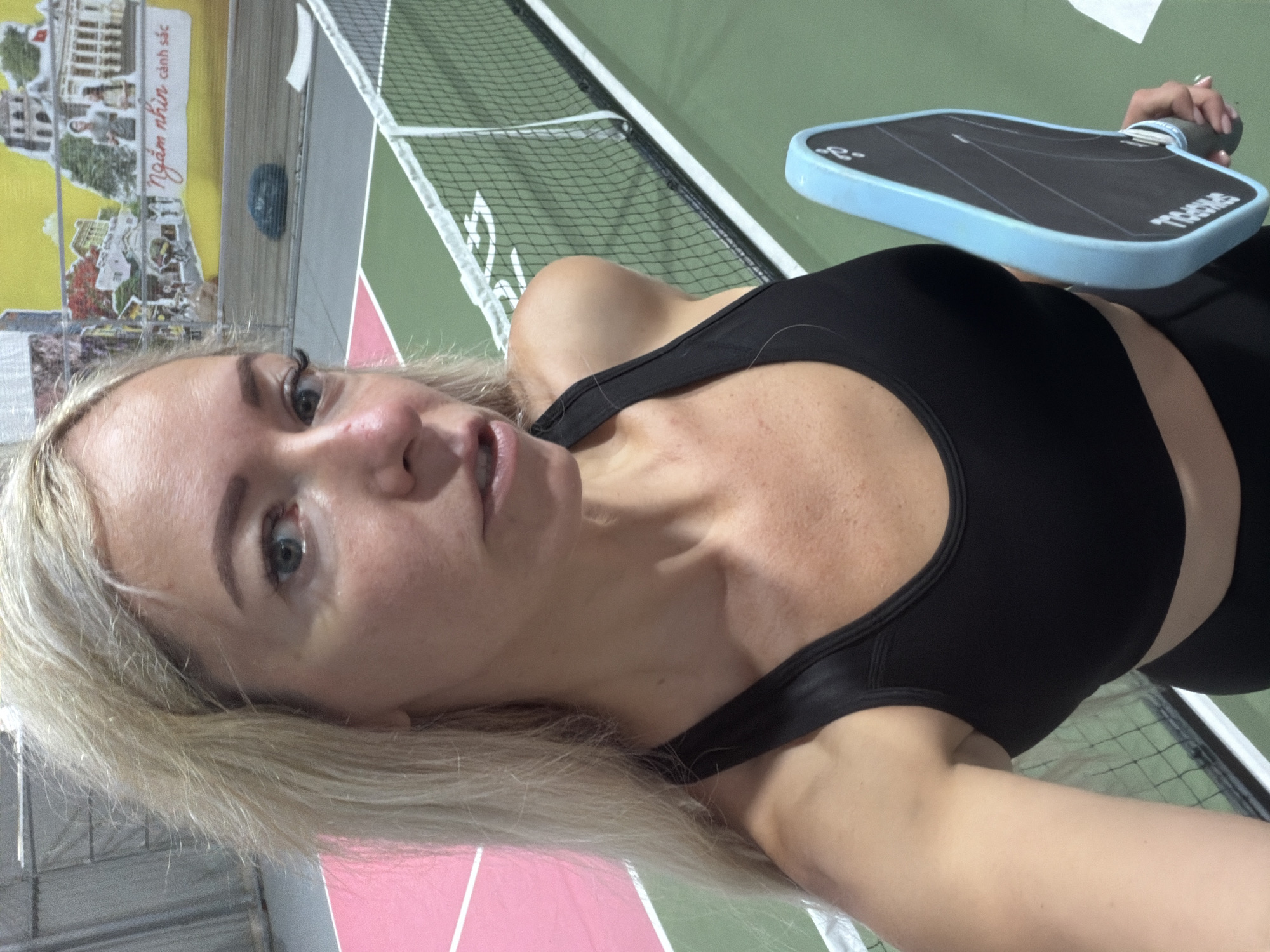 A blonde woman stands on an indoor pickleball court in Da Lat, Vietnam, holding a pickleball paddle. She wears a black sports bra and black leggings. The court has green and pink surfaces, separated by a white line, and a net runs across the middle. In the background, a mural depicts scenes of Vietnamese life, including a building and people.