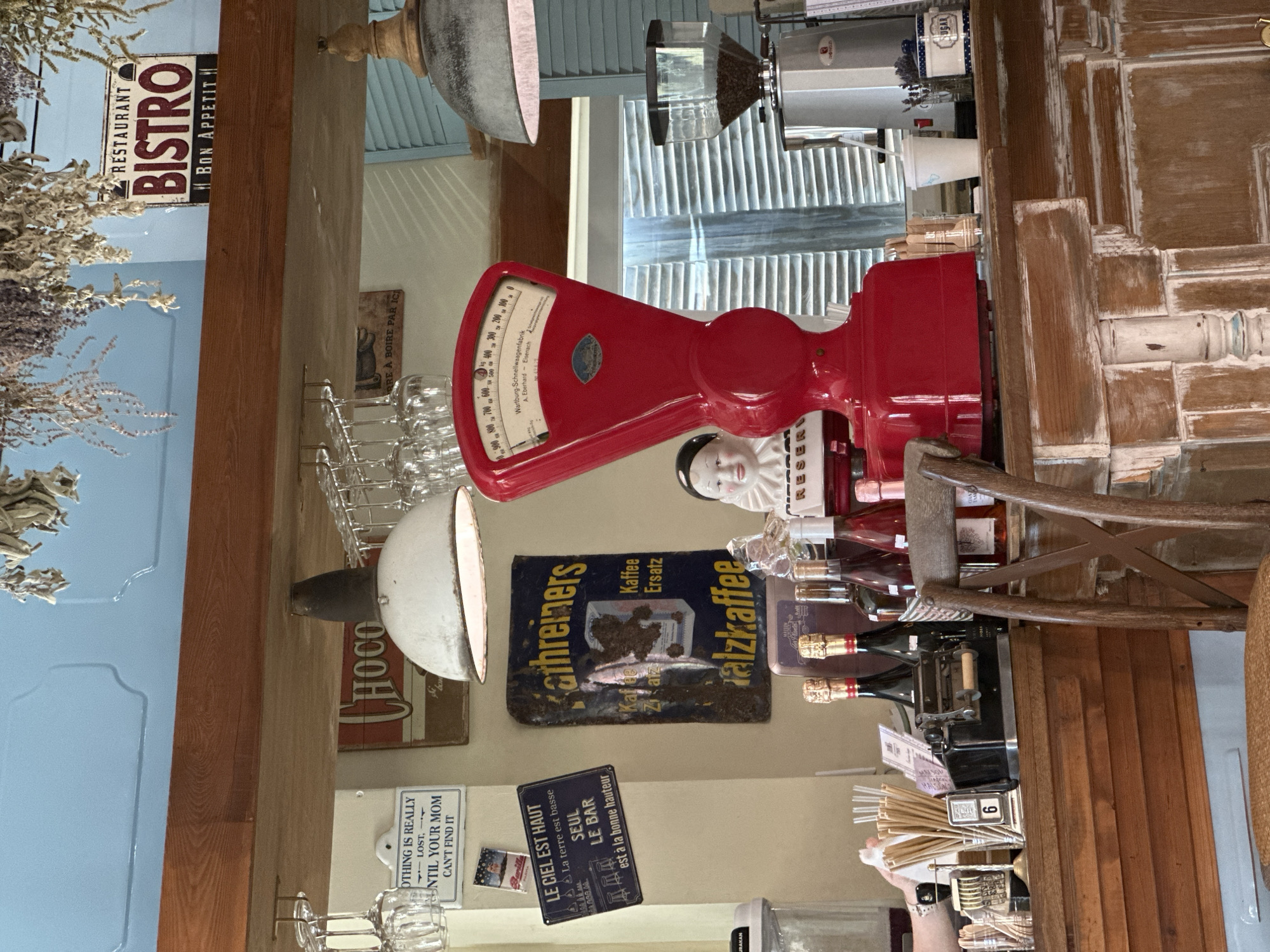 The image depicts an indoor scene of what appears to be a cafe or restaurant counter, likely in Natukhaevskaya, Russia. The foreground features a wooden counter with several items arranged on it. Prominently displayed is a vintage red scale with a circular dial and a platform. To the left of the scale is a porcelain doll with a ruffled collar. Behind the scale, a coffee grinder with a hopper full of beans is visible, along with a white disposable cup and a small jar containing dried lavender.
The background reveals a light blue wall and wooden beams supporting the ceiling. Several decorative signs are affixed to the wall. One sign reads "RESTAURANT BISTRO BON APPETIT". Another sign, with dark blue lettering on a yellow background, advertises "kathreiners SALZKAFFEE". Other signs, including one with French text and a depiction of bar stools, are also present.
On the counter to the left, a collection of paper straws is in a holder, alongside a vintage cash register or receipt imprinter and a small sign displaying the number "6". Several bottles of champagne or sparkling wine, with gold foil around their necks, are lined up. A wooden chair with an "X" shaped back is positioned in front of the counter, partially obscuring the view of the counter's base. The overall impression is one of a cozy, retro-themed establishment. There is no discernible activity or interaction occurring among people in the frame.