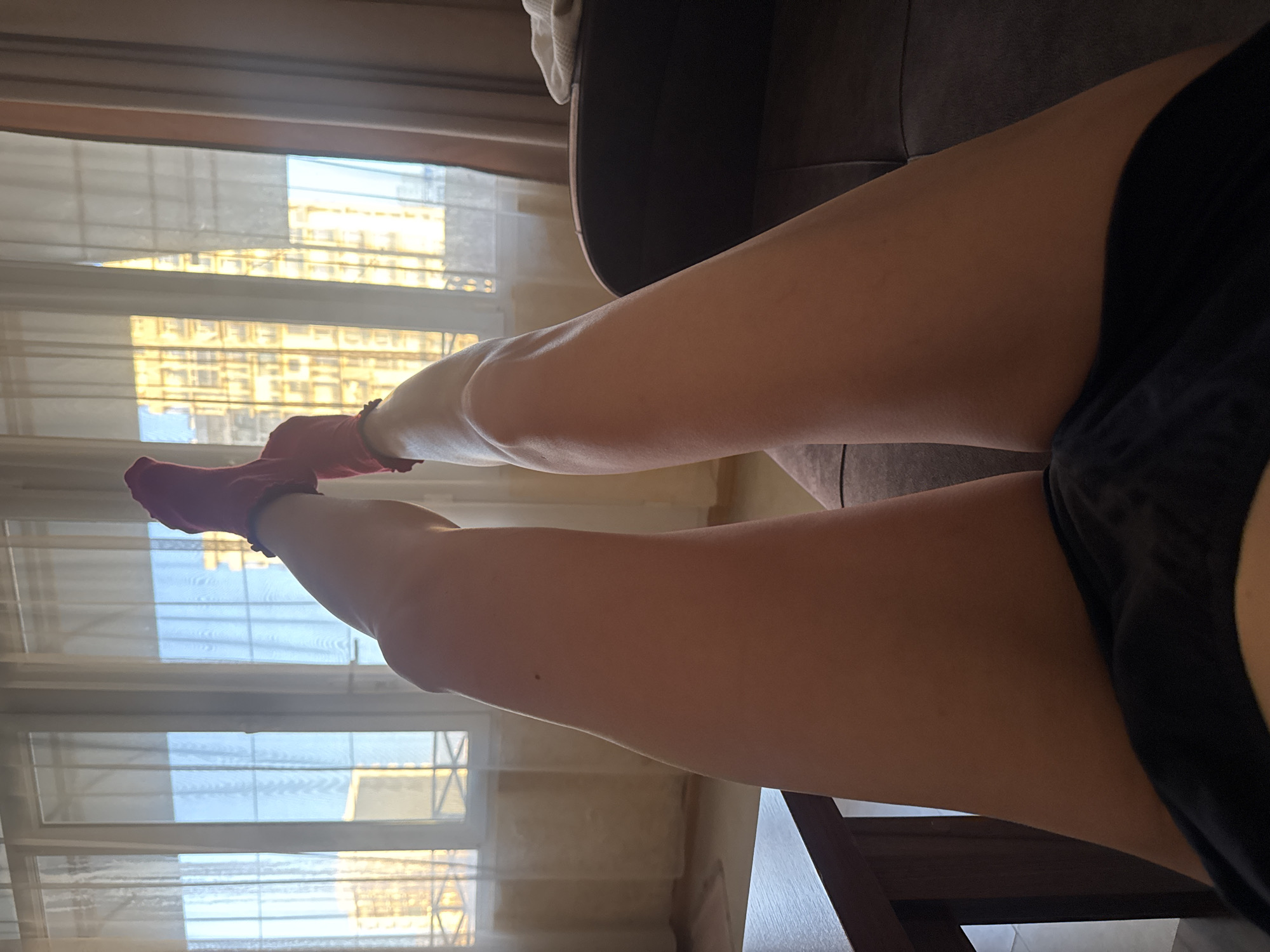 An interior scene is depicted from a low, first-person perspective, focusing on a person's elevated lower body. The subject's bare legs, from mid-thigh to the ankles, are extended upwards and forwards. Both feet are clad in dark red socks. A dark, possibly beaded, anklet is visible on the left ankle. The lower abdomen and upper thighs are partially obscured by dark-colored shorts or underwear.
The legs rest on a dark brown upholstered surface, likely a sofa, visible on the right side of the frame, with a white, folded textile resting on an armrest. To the left, a dark wooden table or furniture piece is partially visible.
The background features a large window with white frames, covered by sheer, translucent light-colored curtains. Through the window, a clear blue sky is visible above an urban landscape. Multiple multi-story buildings, appearing light-colored (possibly beige or yellow) and featuring numerous windows, are illuminated by direct sunlight, indicating daytime. Faint horizontal and vertical lines, suggestive of window bars or balcony railings, are discernible through the sheer curtains in the lower window sections. The overall lighting is bright and natural. The location is an indoor room in a city in Unknown, Unknown.