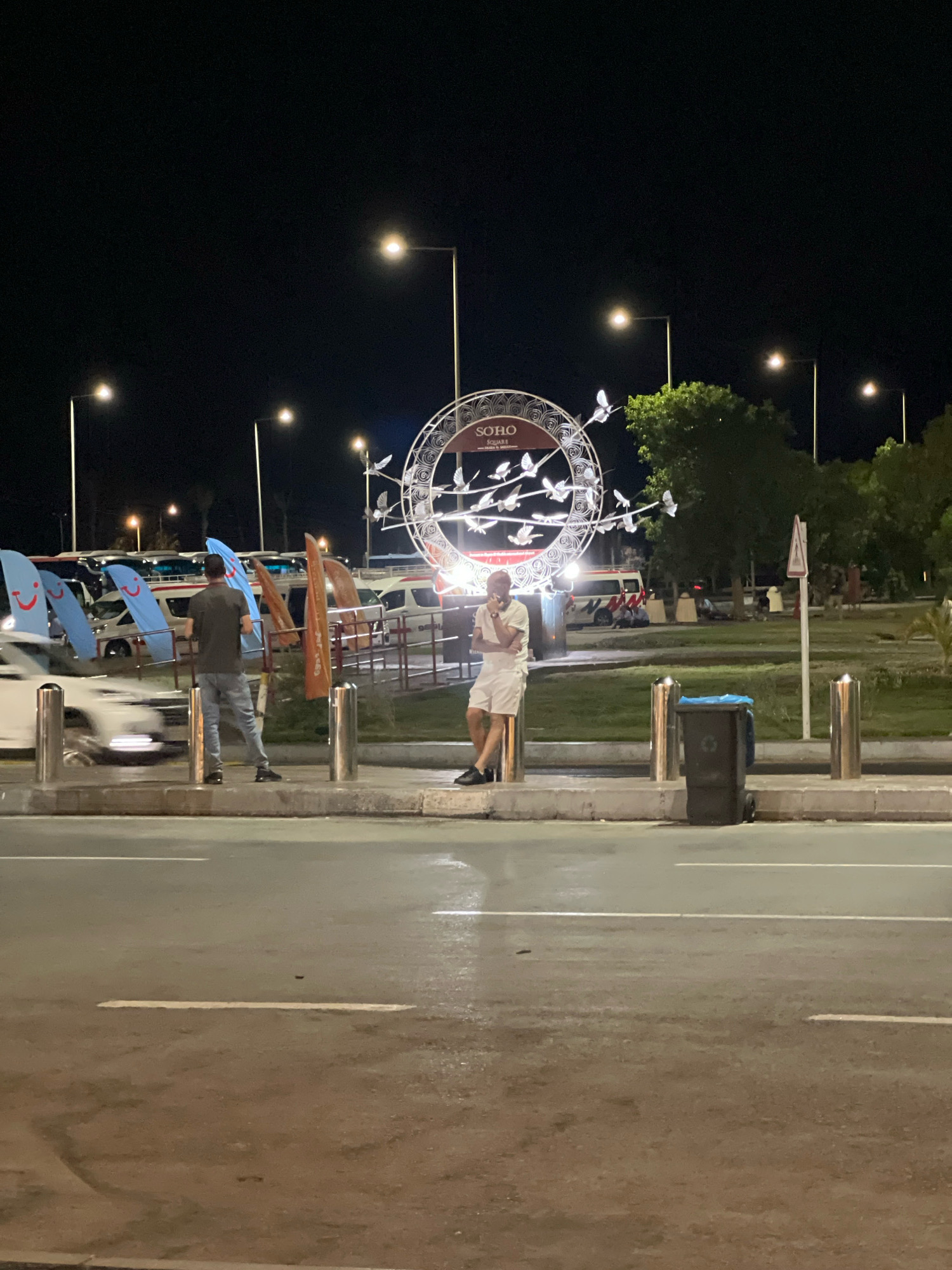 This is a nighttime photograph taken in Sharm El Sheikh, Egypt, likely at Soho Square, given the prominent "SOHO SQUARE" signage on a circular decorative structure. The scene depicts a street or promenade area with several individuals present.
In the foreground, a wet asphalt road with white lane markings is visible. To the left, a white car is blurred due to motion. Behind the car, a person in jeans and a dark t-shirt stands facing away from the camera, near a row of upright metal bollards. To the right of the first person, another individual in white shorts and a light-colored shirt stands leaning against a metal bollard.
Dominating the midground is the large circular decorative structure with the "SOHO SQUARE" text. It is illuminated and features a design of white birds in flight. Behind this structure, there are trees and further background elements suggesting a public space. In the background, there are parked vehicles, including what appear to be vans or minibuses, some adorned with blue flags featuring yellow smiley faces. One orange flag is also visible.
Streetlights are present throughout the scene, casting illumination on the area. A dark trash can with wheels is positioned near the metal bollards on the right side of the frame. The overall lighting indicates it is nighttime, with artificial lights providing the primary illumination. The presence of wet roads suggests recent rain or street cleaning.
