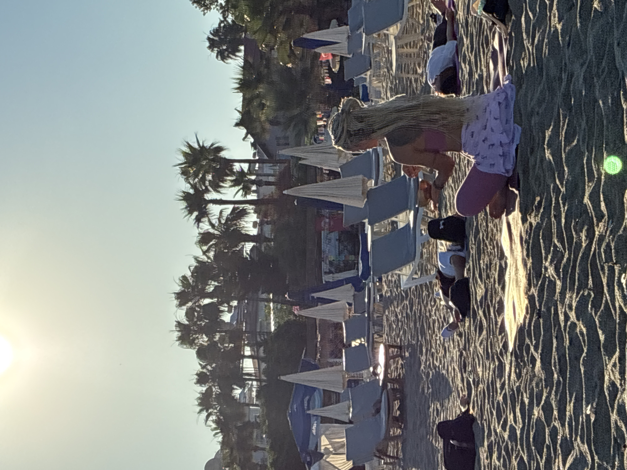 This image depicts a beach scene in Alanya, Turkey, with several people engaged in a yoga or meditation session. In the foreground, a woman with long blonde dreadlocks is seated in a cross-legged position, facing away from the camera. She is wearing a pink top and a light-colored skirt with a patterned design. She appears to be holding a small bowl in her hands, and her posture suggests she is actively participating in the session.
In the mid-ground, several other individuals are lying on yoga mats on the sandy beach. Some are lying on their backs, while others appear to be resting. They are dressed in casual clothing. The beach is lined with rows of empty lounge chairs and beach umbrellas, some of which are open, indicating a resort or public beach setting. The sand is rippled, suggesting the presence of a gentle breeze or the tide.
In the background, palm trees and the architecture of buildings are visible, characteristic of a coastal resort town like Alanya. The sun is high in the sky, casting a bright light and creating some glare, particularly in the upper left corner of the image. The overall atmosphere is peaceful and serene, with a focus on wellness and relaxation.