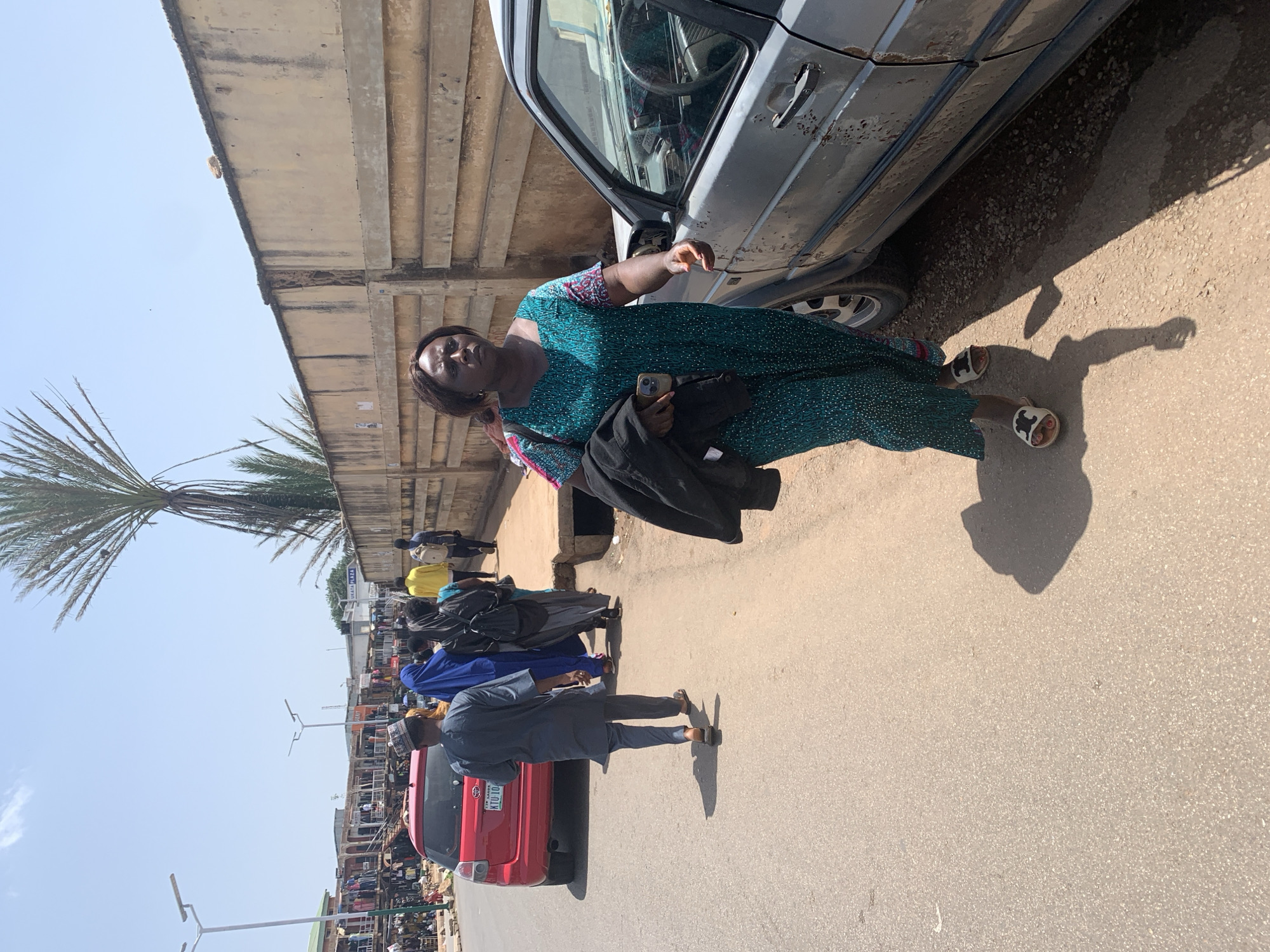 In Jos, Nigeria, a woman in a green, patterned dress walks on a dirt and asphalt road. She carries a black jacket over her left arm and a smartphone in her right hand. Her white sandals have black accents. To her left, a grey car with rust spots is parked on the roadside. Behind her, a group of people walk on the sidewalk, including a man in a grey traditional outfit, a woman in a blue hijab, and another woman in a black hijab carrying a backpack. Further back, a red car and market stalls are visible. The sky is clear and blue, with a tall palm tree to the left of the frame and a beige concrete wall along the right side.