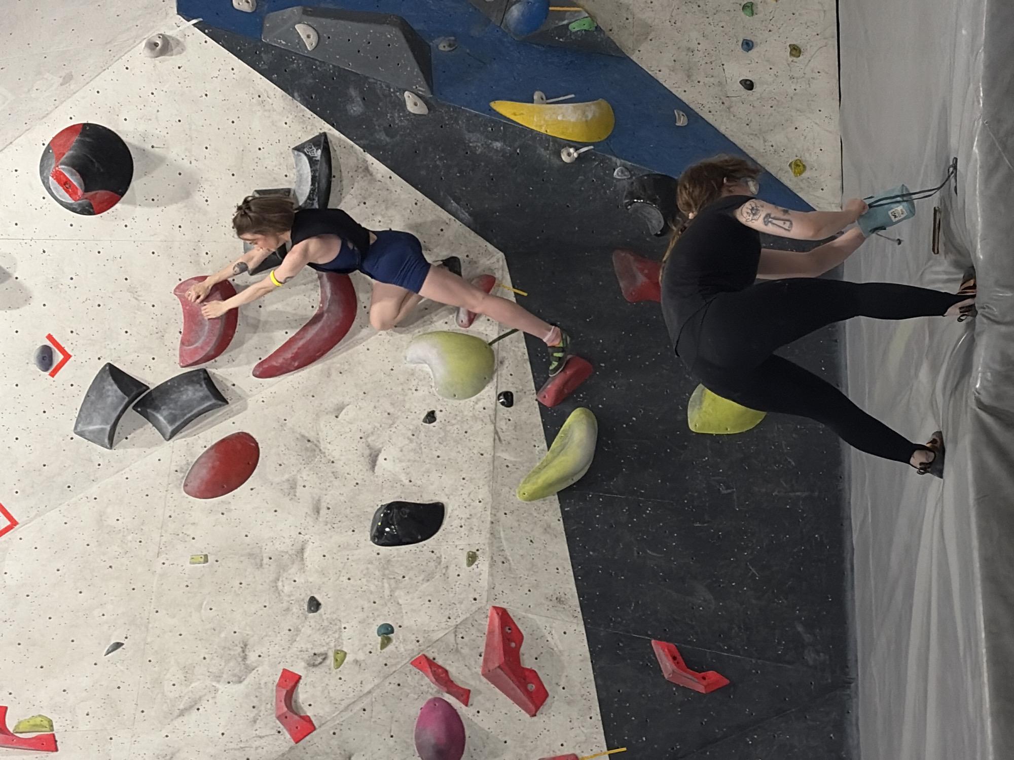 The image depicts an indoor climbing gym in Moscow, Russia. Two individuals are present. In the upper portion of the frame, a woman with short brown hair, wearing a dark blue athletic top and shorts, is actively climbing a textured wall. She is reaching for a red climbing hold with her right hand, her left hand also engaged on a hold. Her feet are positioned on other holds, and she has a yellow wristband on her left wrist. The climbing wall features numerous colorful holds of various shapes and sizes, predominantly red, gray, and yellow, against a white and black patterned background. In the lower portion of the frame, another person, likely a woman based on the visible tattoos on her arm and the styling of her hair, is bent over on a padded mat at the base of the wall. She is wearing black leggings and a black t-shirt and is holding a light blue chalk bag, appearing to prepare to use it. The environment is a controlled indoor space designed for bouldering.