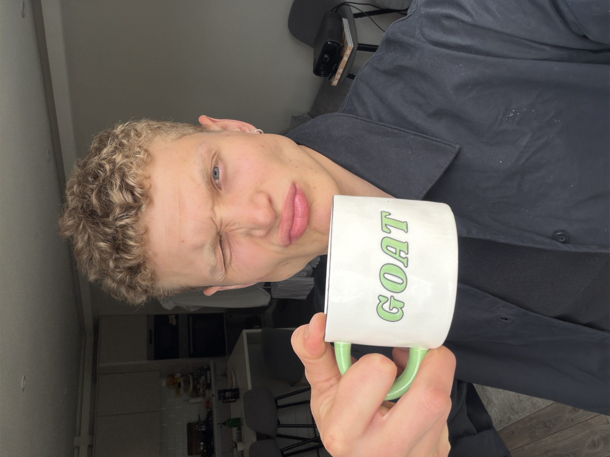 A young adult with curly blonde hair and blue eyes, making a puckered face, holds a white mug with a green handle and the word "GOAT" in green lettering. The person is wearing a dark collared shirt. The background suggests an indoor environment, possibly a kitchen or living area, with cabinets and a table visible. The lighting is bright, and the image appears to be a selfie. The specific location within London is not discernible from the visual evidence.