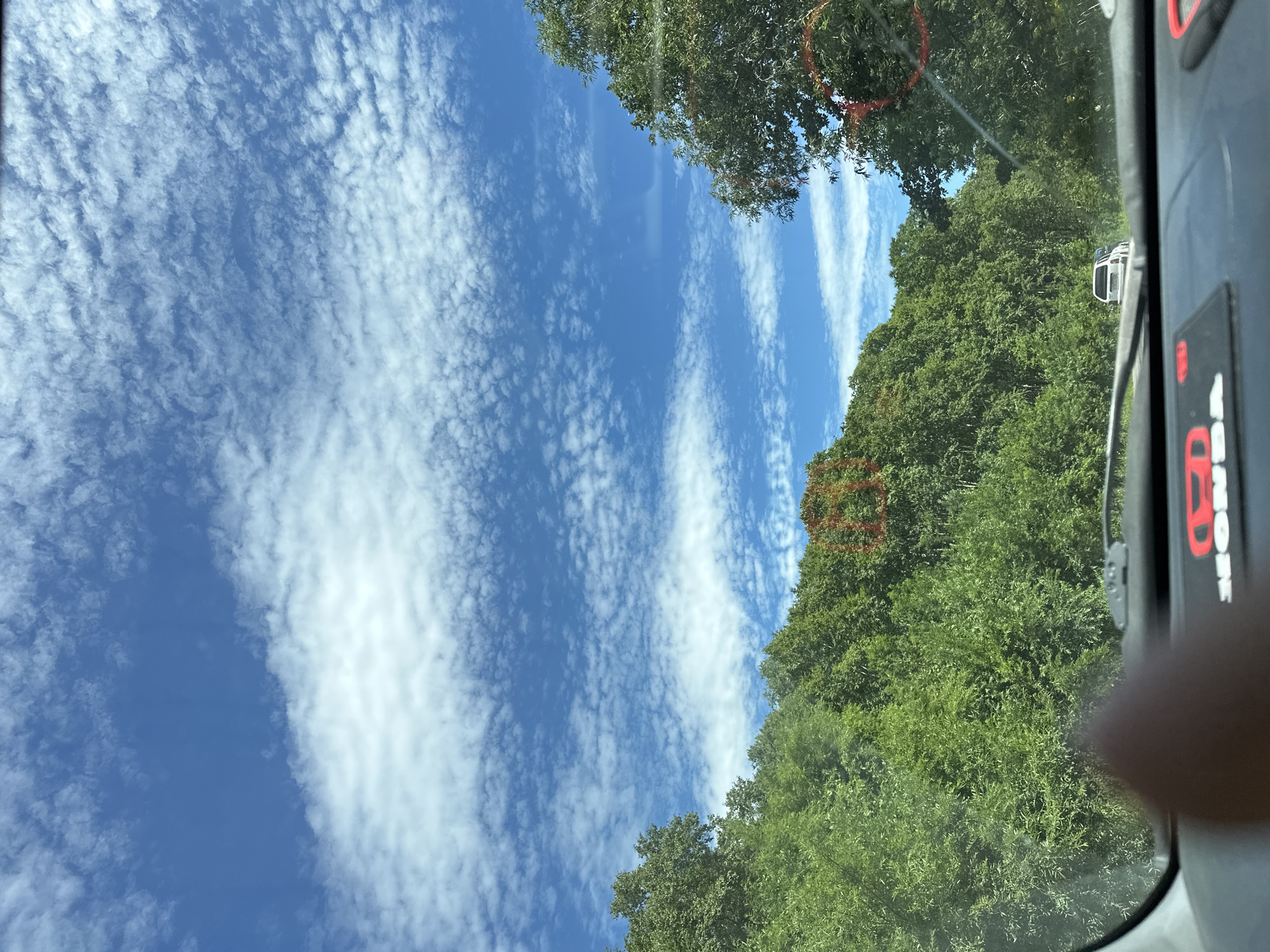 The image shows a view from inside a vehicle, looking out through the windshield. The sky is blue with scattered white clouds. Dense green trees line a road, with a white car visible in the distance. The dashboard of the vehicle is partially visible in the foreground. The image was captured in Petropavlovsk-Kamchatsky, Russia.