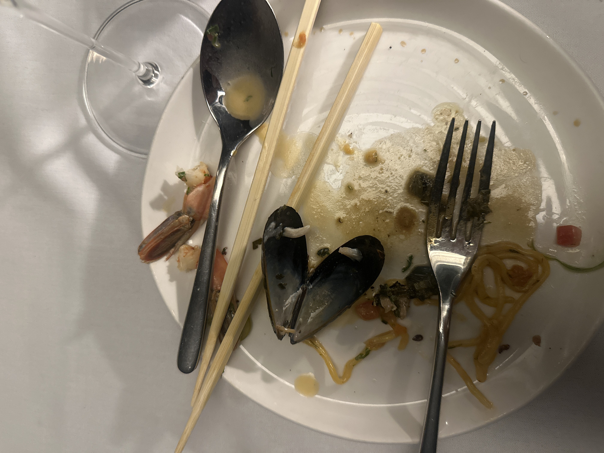 The scene depicts a partially eaten meal on a white plate. The remnants of the meal include a shrimp, mussel shells, pasta, and sauce. Utensils present are a spoon, fork, and chopsticks. A partially visible wine glass sits beside the plate. Given the presence of shellfish and pasta, it appears to be a seafood-based dish. The location is potentially a restaurant in Tel-Aviv, Israel.