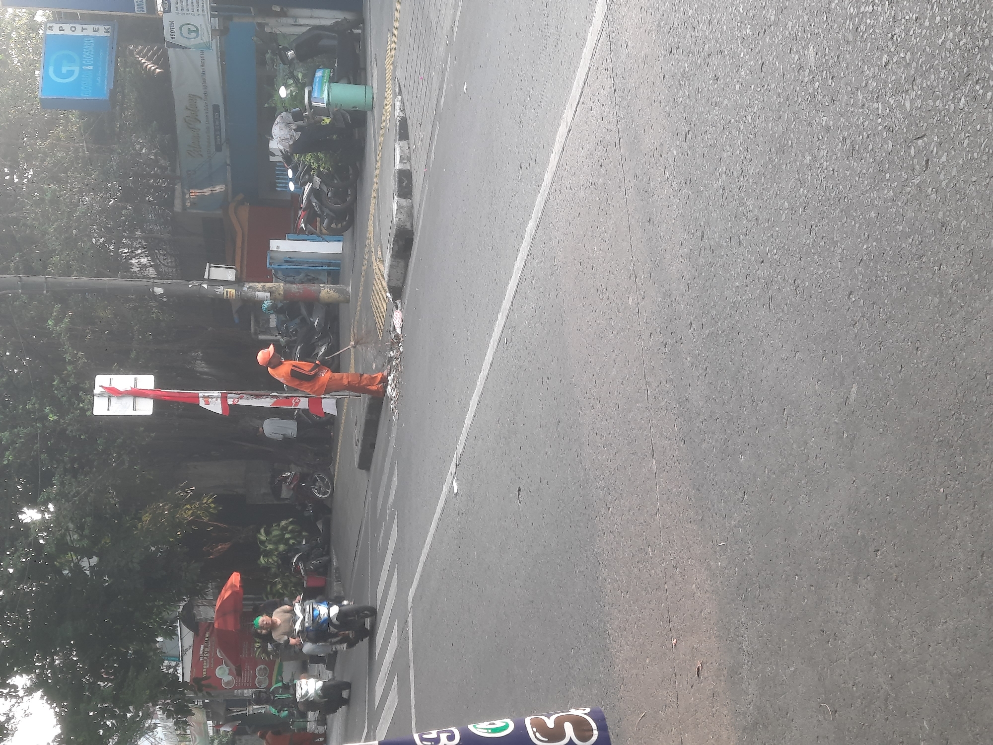 An outdoor, medium-range, eye-level, daytime, street-level shot captures a scene in North Jakarta, Indonesia. The frame is dominated by a grey asphalt road with white lane markings. A pedestrian crosswalk is visible in the mid-ground. To the right of the crosswalk, a sanitation worker wearing an orange uniform and cap is sweeping debris with a broom. The sanitation worker is standing near a green trash receptacle. Behind the sanitation worker and crosswalk, several motorcycles are parked along a sidewalk. The sidewalk is lined with buildings and trees. One building has a blue sign with white lettering that reads "APOTEK". Further back, a red umbrella is set up outside a food stall. Several people are on motorcycles in the background, and a few are visible walking. The sky is overcast and hazy. A blue and white cone is in the foreground on the left.