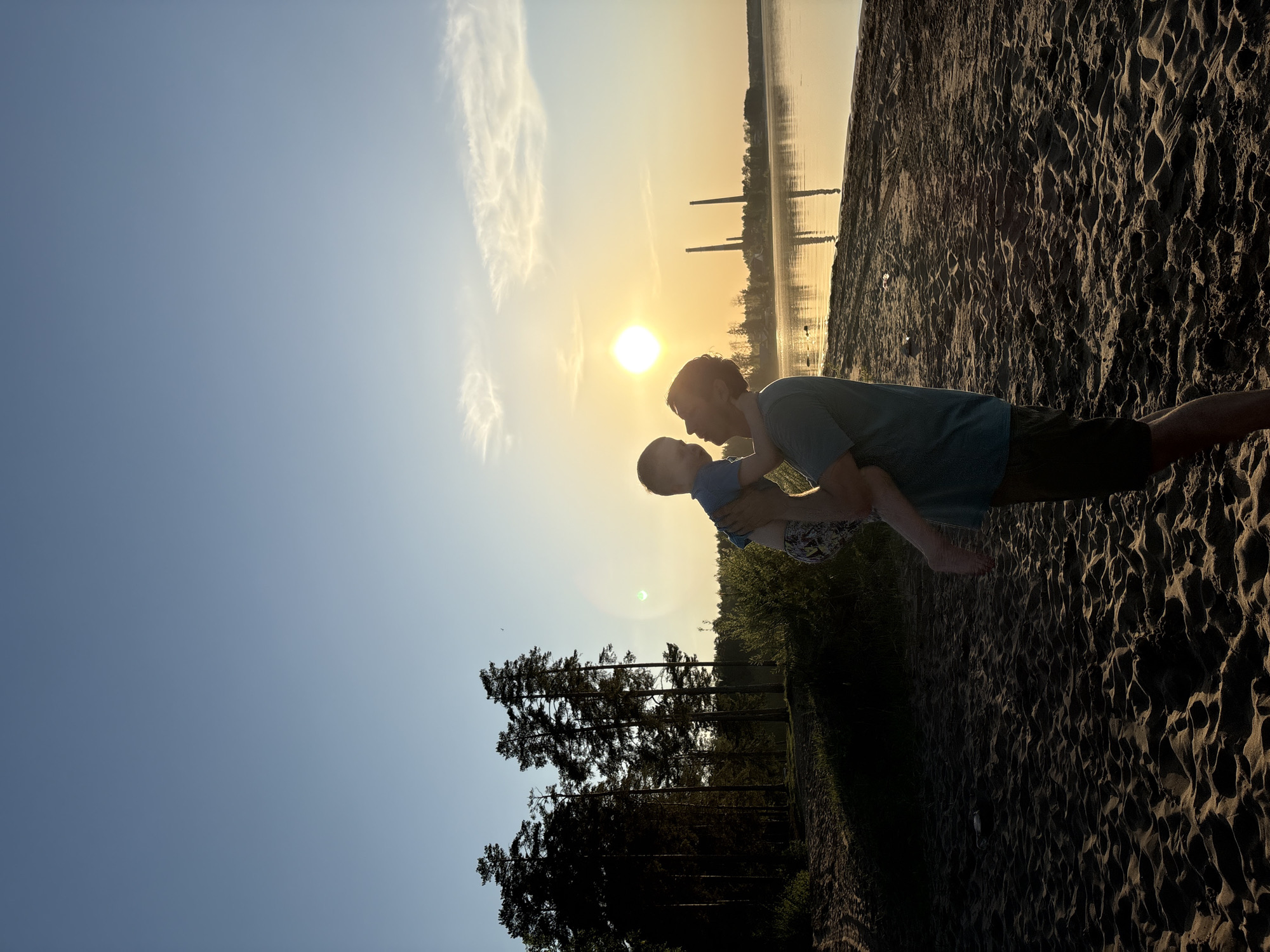 The image shows a man holding a young child, possibly a toddler, against a bright sun. They appear to be on a sandy beach, with trees and vegetation at the bottom of the frame. The background shows a body of water and structures that could be industrial chimneys or towers, possibly indicating a city in the distance. Based on the prompt mentioning Chaikovsky, Russia, the location is likely near a body of water there.