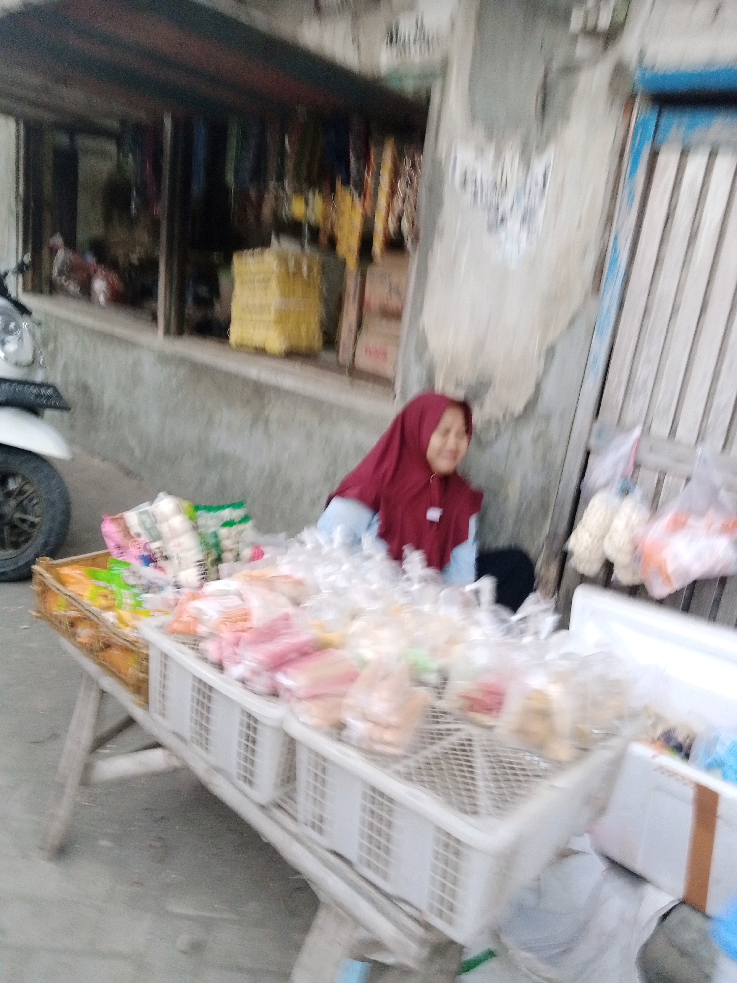 An outdoor market stall in Sono Ageng, Indonesia, features a vendor wearing a red hijab and blue shirt, serving a customer who is out of frame. The stall displays a variety of packaged snacks and food items in clear plastic bags, arranged in white plastic crates on a wooden table. Behind the stall, a building with a weathered exterior and a metal shuttered door is visible, with some shelves stocked with goods. A motorcycle is parked to the left of the scene. The ground is paved with a gray surface.