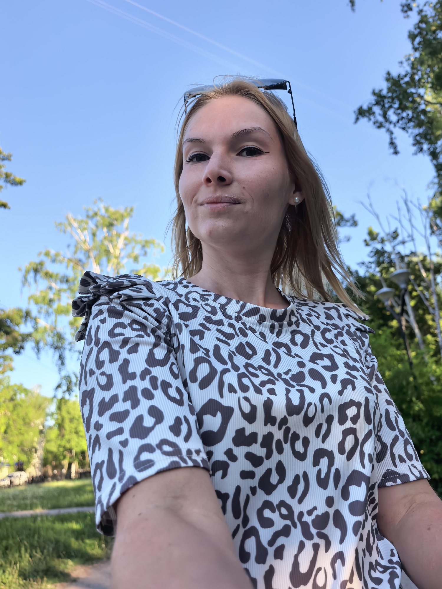 The image is a low-angle, close-up selfie of a woman with fair skin, blonde hair, and light-colored eyes. She is wearing a cream-colored top with a brown leopard print and a ruffled shoulder detail. Sunglasses are perched on her head.
The background features a blue sky with faint contrails. Lush green trees and foliage are visible. It is daytime, and the lighting is bright.
Given the prompt constraints, specifying the precise location in Sterlitamak, Russia, is not possible without additional information or features identifiable to that specific location. The background shows a general park or green space environment.