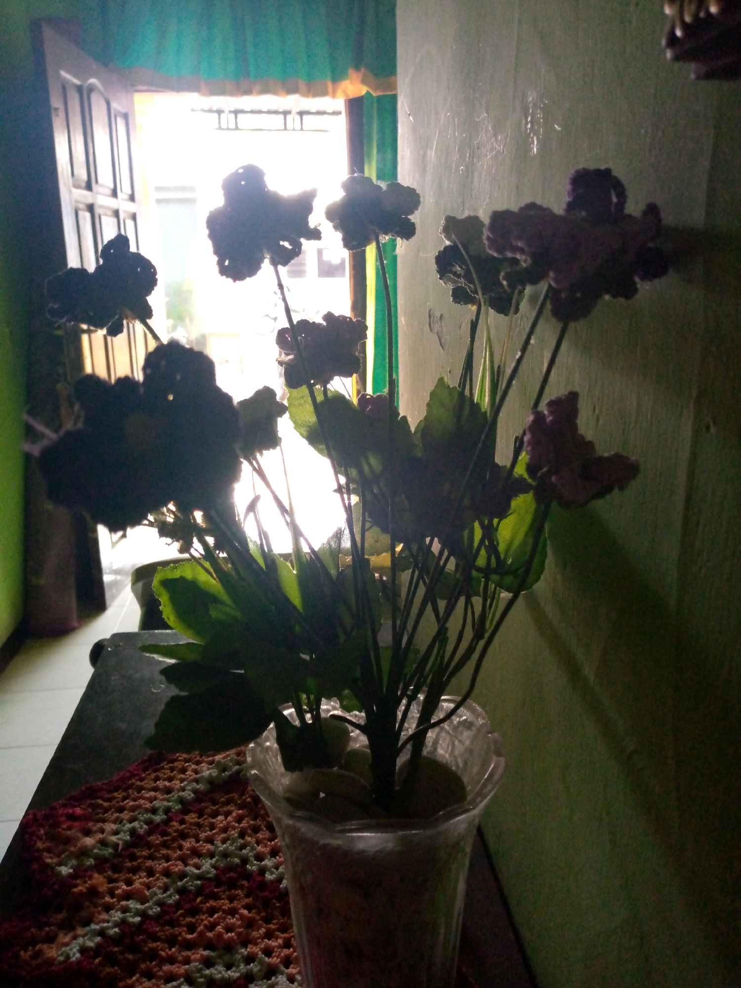 The image shows a potted artificial flower arrangement on a surface, possibly a table or shelf, in an indoor setting with a doorway or window in the background. The flowers, which appear dark and clustered, are silhouetted against bright natural light coming from the open doorway. The wall behind the flowers is a textured green. In the foreground, a crocheted mat with a pattern of red, orange, and white is partially visible. The overall lighting suggests the image was taken during the day, possibly late afternoon, given the strong backlight. The location is described as Sumberwono, Indonesia. There are no people visible in the image, and the focus is on the still life arrangement. The room appears to be simply furnished, with the green wall and doorway being the most prominent environmental features aside from the flowers.