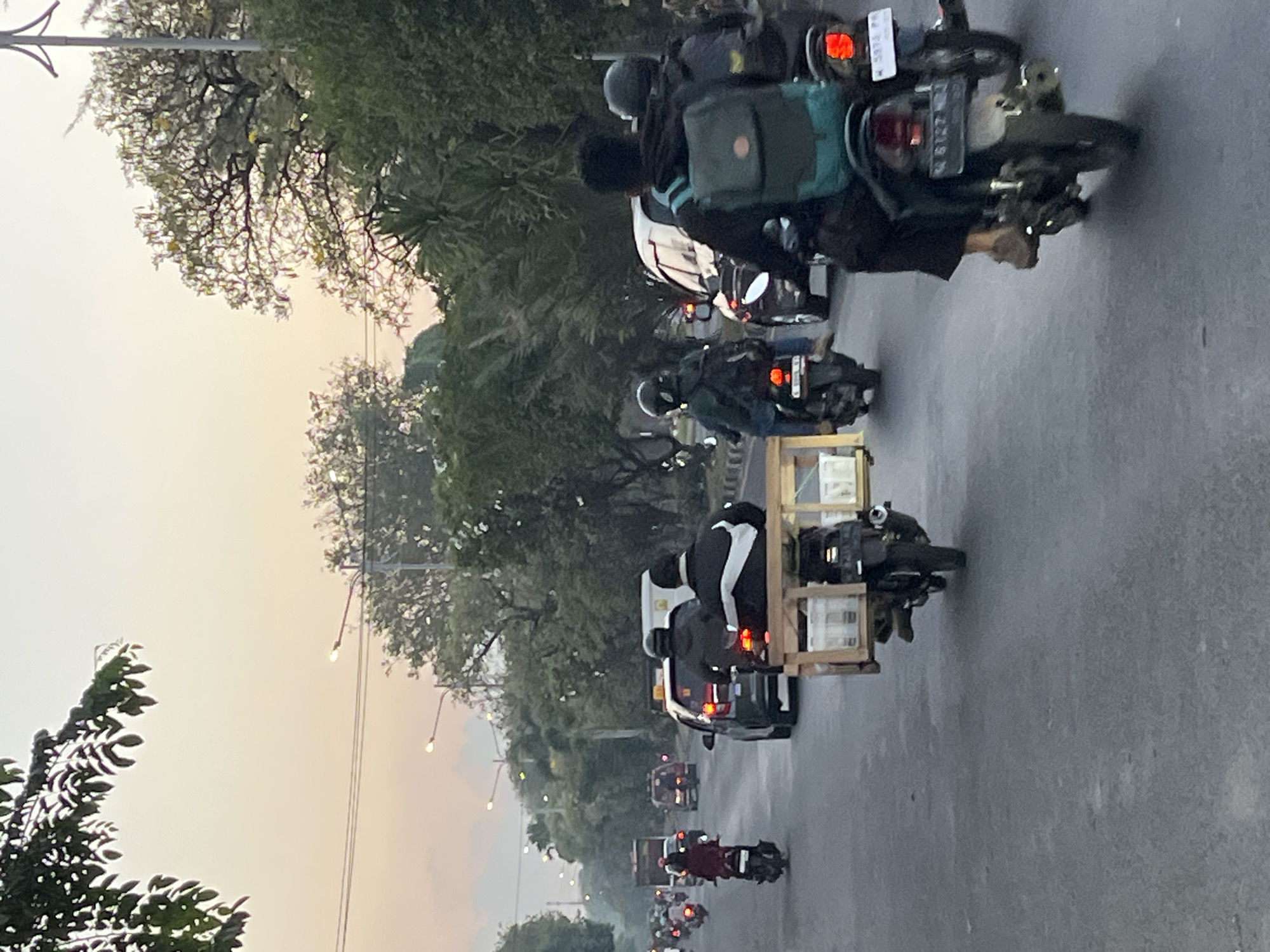 The image shows a street scene in Ngaban, Indonesia, with several motorcycles and cars on a road. The sky is overcast with a pale orange hue, suggesting it might be dawn or dusk. Trees line the sides of the road, and streetlights are visible in the distance.
In the foreground, a motorcycle is carrying a wooden crate filled with bottles. Two people are riding on this motorcycle, both wearing jackets. The rider in the back is wearing a dark jacket with a white stripe on the back, and the passenger in front is wearing a black jacket.
Further down the road, other motorcycles and cars are visible. One car is dark-colored and appears to be a sedan. Another motorcycle has two riders, both wearing helmets and jackets. A motorcycle in the middle ground has a single rider wearing a red jacket. In the far distance, more vehicles are present, and some buildings or structures can be seen. The road surface is asphalt and appears to be wet in some areas, possibly from recent rain. The overall impression is of a busy street during a transitional time of day.