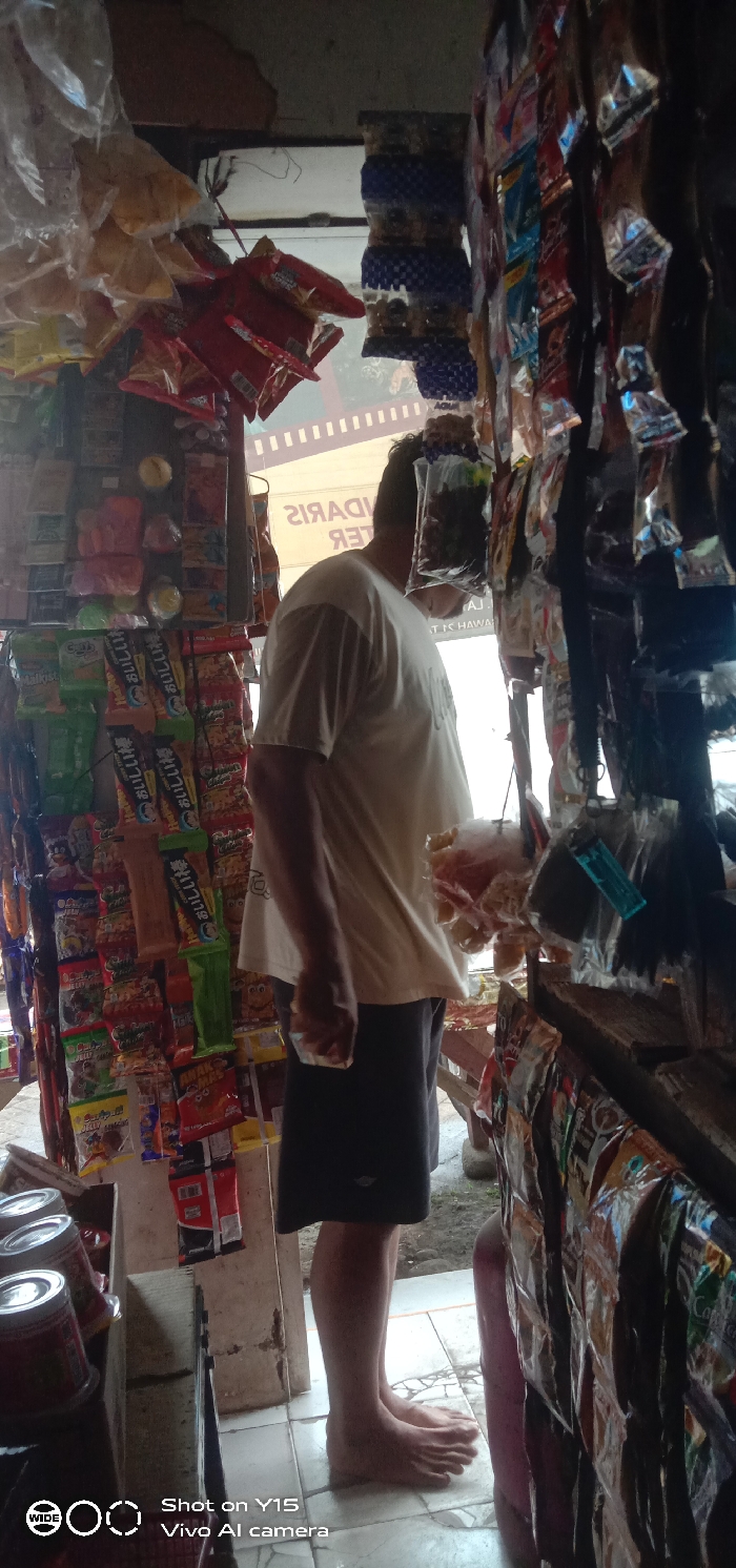 An interior view depicts a small, densely packed convenience store, or "warung," located in Sebaung, Indonesia. The scene features a single adult male, viewed from the upper back and right side, standing barefoot on light-colored tiled flooring. He is wearing a light-colored, short-sleeved t-shirt and dark shorts, with his head slightly bowed and turned to the right.
The shop's interior is dominated by an extensive display of packaged goods. On the left, a vertical rack is laden with various snacks, candies, and instant food items, including multi-colored bags (green, red, orange) and what appear to be instant noodles. Below these, several red-labeled cylindrical cans are stacked. The right side of the image features a curtain-like arrangement of numerous individually packaged items, some with blue and white checkered patterns, hanging vertically from overhead structures. Additional items in clear plastic bags are suspended around the male's head level. A red plastic container is partially visible on the far right foreground.
Bright natural light emanates from an opening behind the individual, illuminating an exterior space that includes visible ground and a partially legible, inverted sign displaying Indonesian text like "TER" and "MARIS," possibly part of a shop name. The shop's structure appears to consist of simple shelving or wooden supports for the merchandise. The overall impression is that of a bustling, local retail establishment.