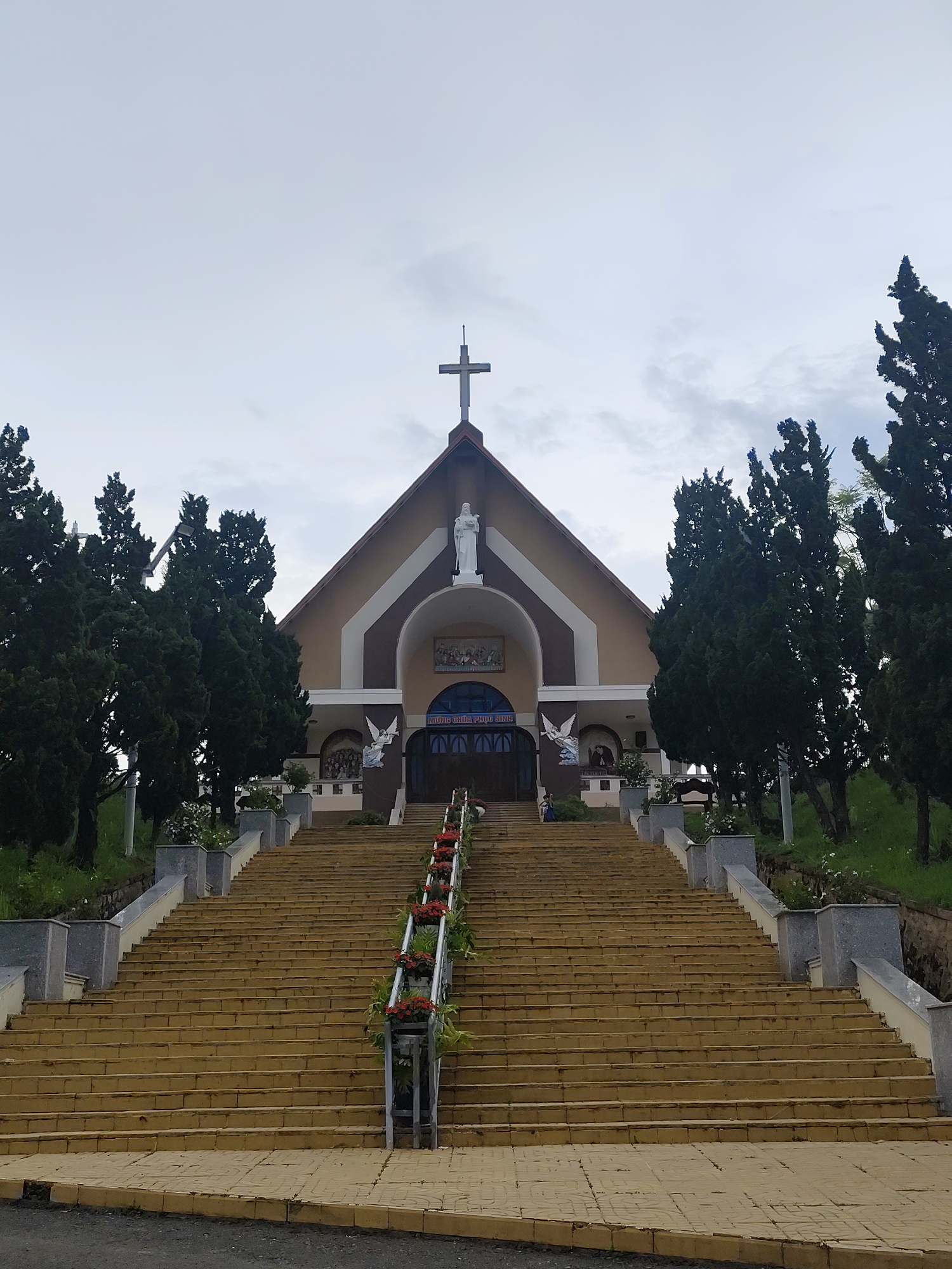 A low-angle, eye-level shot depicts a large church, accessed by a lengthy staircase in Lạc Dương, Vietnam.
The church is a light brown color with dark brown accents, featuring a triangular roof with a large Christian cross at its peak. A white statue is positioned within an archway below the cross. The facade includes arched windows above the entrance doors, which are dark blue. Angelic figures are visible on either side of the doors.
A long, wide staircase leads up to the church entrance. The stairs are light brown with gray accents and are flanked by trees and flower planters. Flower boxes with red and green plants line the staircase.
The sky is overcast and gray. The scene is mostly static. A few people can be seen at the top of the staircase. The surrounding environment includes trees and landscaping. The road is paved and located at the bottom of the stairs.