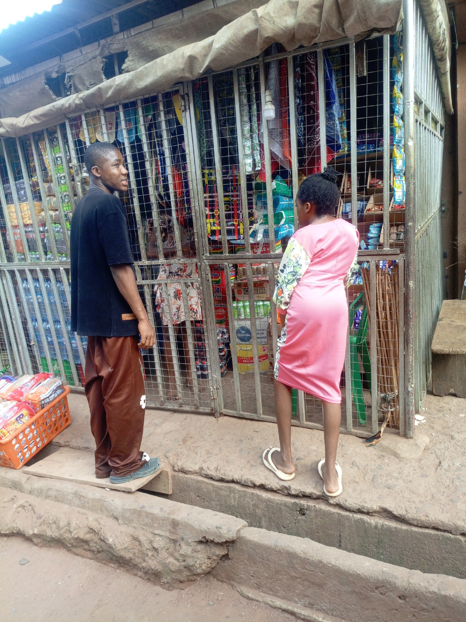 An outdoor scene in Onitsha, Nigeria, shows two individuals interacting with a small, informal shop or kiosk. The shop is secured by a tall, gray metal grid gate, behind which shelves are packed with various consumer goods, including bottled drinks (some appearing to be Sprite), packaged snacks, and other items. A tattered, light-colored canvas awning covers the top of the shop.
On the left, a young man with short, dark hair stands, looking towards the left with a slight smile. He is wearing a black short-sleeved t-shirt with a faint "LOADED" label visible near the hem, and baggy brown pants. He stands on a wooden plank that bridges a concrete gutter or drainage ditch in the foreground. To his left, an orange plastic basket filled with packaged bread or similar goods rests on the ground.
On the right, a woman stands with her back to the viewer, facing the shop. She is wearing a pink knee-length dress with patterned sleeves and white flip-flops. Her hair is tied up.
The ground is unpaved and uneven, suggesting a typical informal market or street setting. Natural daylight indicates it is daytime. The scene depicts a casual moment of daily commerce in a local urban environment.