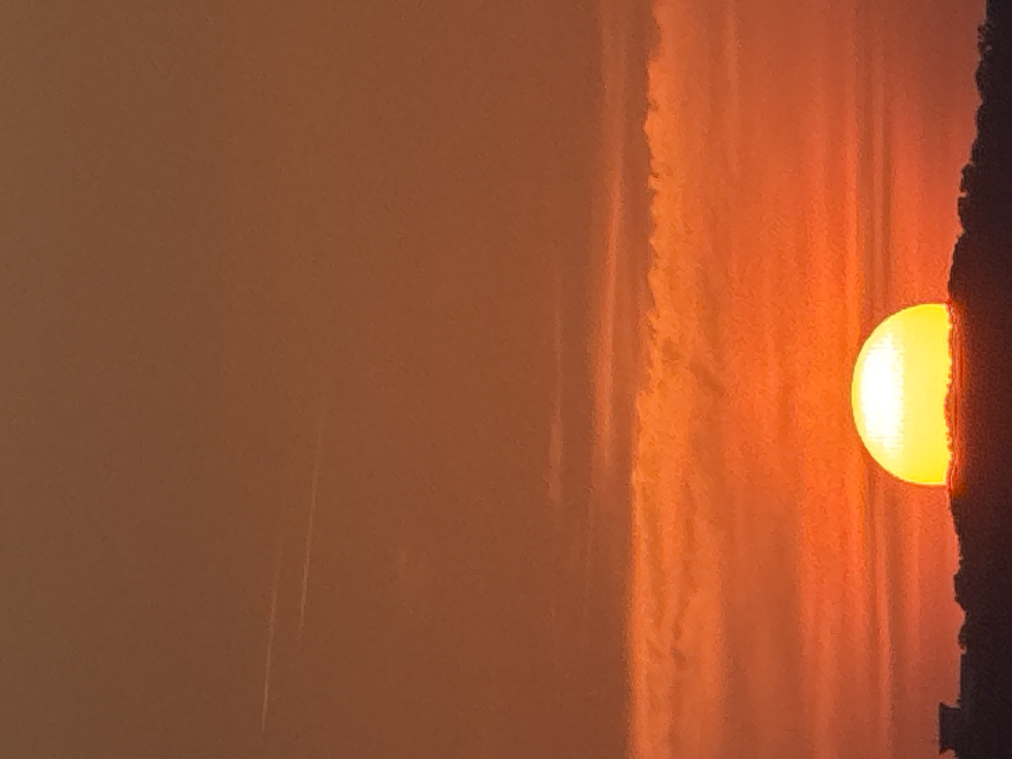 The image shows a sunset over a silhouetted landscape of buildings and trees. The sun, a brilliant yellow sphere, is partially obscured by the horizon, casting an orange glow across the sky. Wispy clouds are visible in the distance, and two faint contrails from an aircraft stretch across the upper portion of the sky. The overall impression is one of a peaceful evening scene.