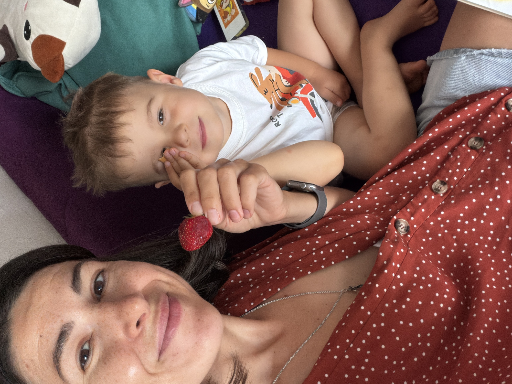 A high-angle selfie captures an intimate moment between an adult woman and a young child relaxing indoors. The woman is in the foreground, lying down and looking towards the camera with a warm, gentle smile. She has dark hair and freckles, wearing a reddish-brown polka dot top with buttons and a silver necklace. A grey smartwatch is visible on her left wrist. She holds a ripe red strawberry delicately in her fingers, positioned near her face.
Behind her, to the left, a young child, likely a boy, is also lying down and gazing towards the camera with a soft smile. He has light brown hair and is dressed in a white t-shirt featuring a colorful graphic, where the partial text "ROY" is visible. A white and brown plush toy, resembling a cow, rests above his head. Colorful cards or books, one showing an image similar to Paw Patrol characters, are visible in the background near the child.
The setting appears to be a cozy indoor space, likely a sofa or bed, indicated by the soft purple and dark green upholstery. The overall atmosphere conveys relaxation and affection. The image was taken in Budva, Montenegro, during what appears to be daytime.
