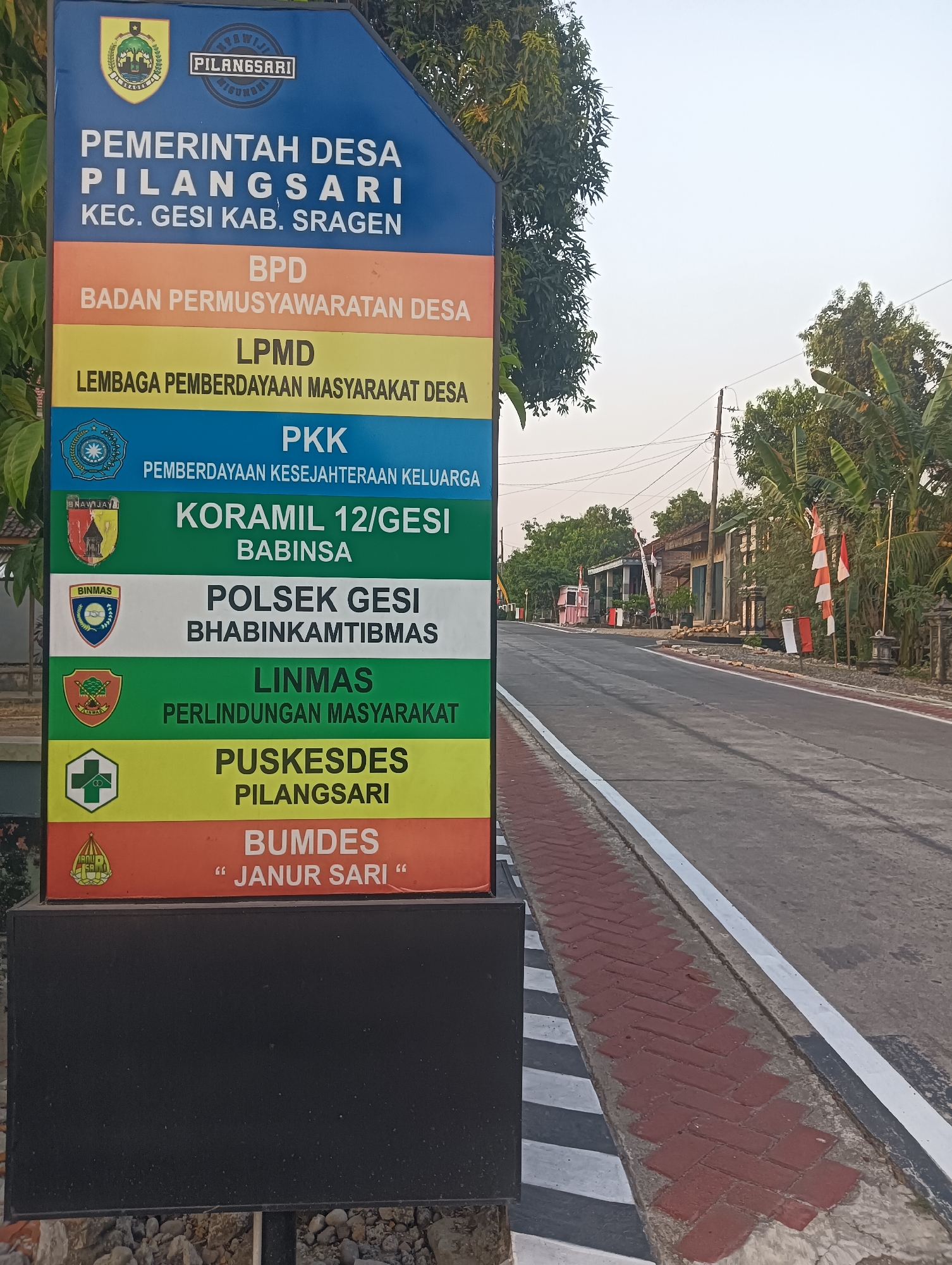 A vertical, multi-colored sign stands on the left side of a paved road in the Indonesian countryside. The sign, with a blue top section, lists various government and community organizations of Pilangsari village, Gesi sub-district, Sragen regency. These include "PEMERINTAH DESA PILANGSARI", "BPD BADAN PERMUSYAWARATAN DESA", "LPMD LEMBAGA PEMBERDAYAAN MASYARAKAT DESA", "PKK PEMBERDAYAAN KESEJAHTERAAN KELUARGA", "KORAMIL 12/GESI BABINSA", "POLSEK GESI BHABINKAMTIBMAS", "LINMAS PERLINDUNGAN MASYARAKAT", "PUSKESDES PILANGSARI", and "BUMDES "JANUR SARI"". The sign is mounted on a black base. The road curves gently to the right and is bordered by a red brick walkway on the left. In the background, houses, trees, and power lines are visible under a clear, bright sky. Several Indonesian flags are also displayed on the right side of the road. There are no people visible in the image. The scene is likely set in the village of Pilangsari, Gesi sub-district, Sragen regency, Central Java, Indonesia.