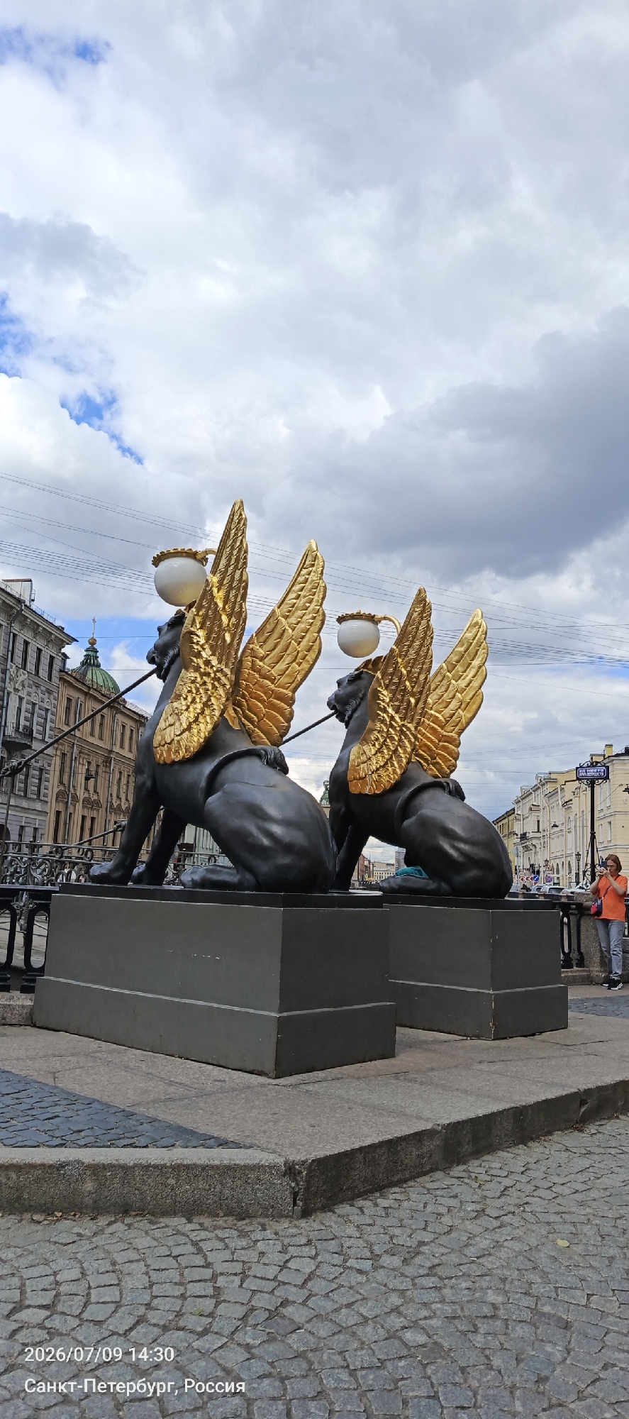 The image shows two sphinx sculptures with golden wings, each supporting a lamp with a white spherical shade. The sculptures are black and sit on dark grey rectangular pedestals. The pedestals are placed on a cobbled pavement. In the background, there are buildings with classical architecture and a domed building. A woman wearing an orange shirt and gray pants stands on the right, holding a camera. The sky is cloudy with patches of blue. The timestamp and location "2026/07/09 14:30, Санкт-Петербург, Россия" are visible in the bottom left corner, indicating the photograph was taken on July 9, 2026, at 2:30 PM in Saint Petersburg, Russia.