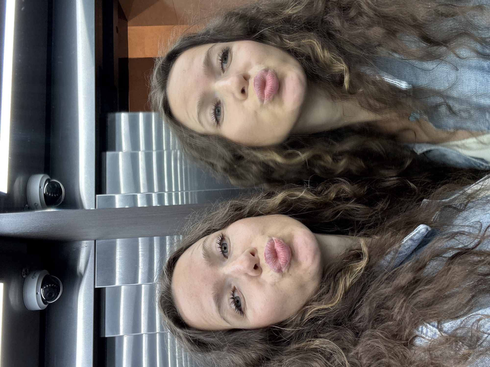 In a close-up selfie taken in an elevator, two young women with long, curly brown hair pose with puckered lips. They are positioned side-by-side, filling the frame. Both women are looking towards the camera. The left woman has fair skin with a few freckles and light blue eyes. The right woman, whose face is slightly less visible, also appears to have fair skin and light blue eyes. They are both wearing light blue denim shirts. In the background, a dark wooden panel divides the elevator from a reflective metal surface with horizontal lines. Above them, two dome-shaped security cameras are visible, mounted on a black ceiling. The lighting suggests an indoor environment, possibly a public building. The overall scene captures a casual and playful moment between two individuals in an urban setting.