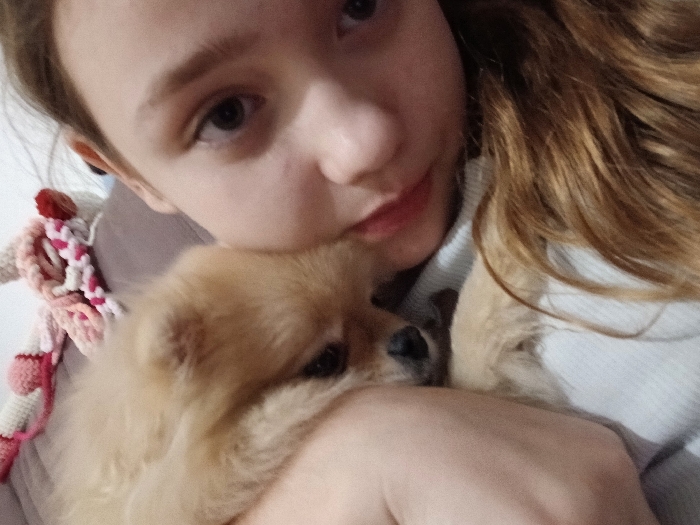 A young girl holds a light brown Pomeranian puppy close. She faces the camera with a soft smile, her gray eyes looking directly at the viewer. Part of a crocheted toy is visible to her left. The girl wears a light-colored, possibly white, shirt. Her hair is light brown, partially in focus. The puppy appears relaxed, nestled against her, its eyes partially closed. The lighting is diffused, creating a gentle, warm tone.