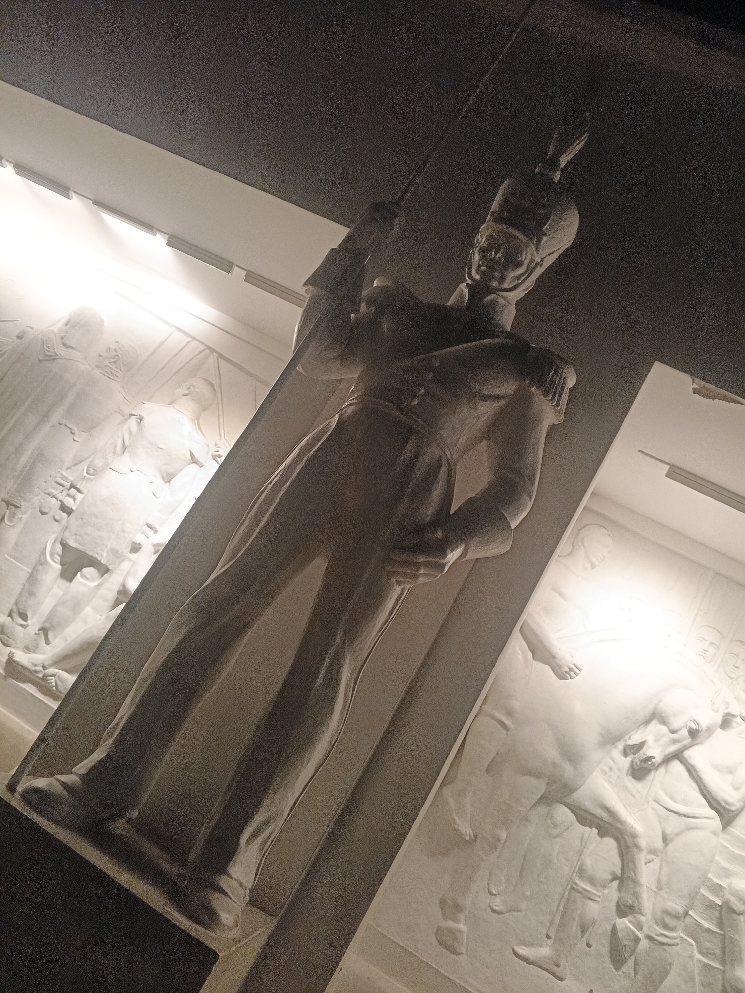 The image shows a sculpted monument of a soldier in uniform holding a spear. The sculpture is positioned in a dimly lit indoor space, with spotlights illuminating the monument and a bas-relief sculpture on the wall behind it. The bas-relief depicts figures on horseback and marching soldiers. The overall impression is one of historical and military commemoration. This monument is located in the Plaza Bolivar in Caracas, Venezuela.