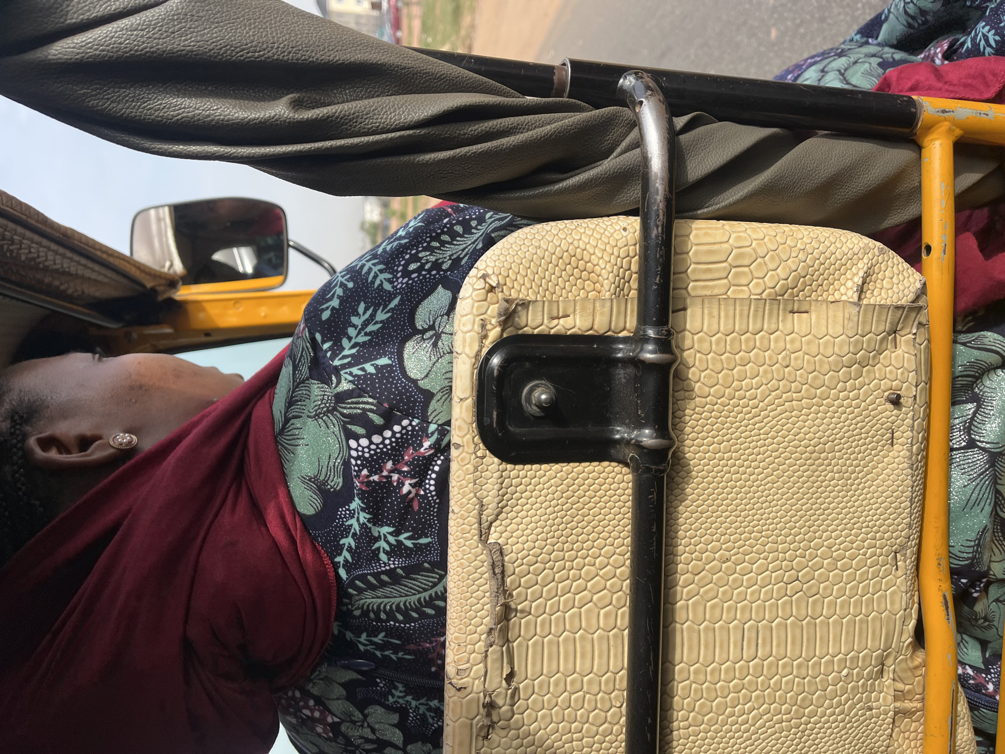 The image shows the interior of a vehicle, possibly an auto rickshaw, in Galadimawa Village, Nigeria. An individual is visible, wearing a dark floral-patterned garment and a red shawl. The vehicle interior features a light-colored, reptile-skin-textured seat. Metal bars and a tan leather canopy are also visible.