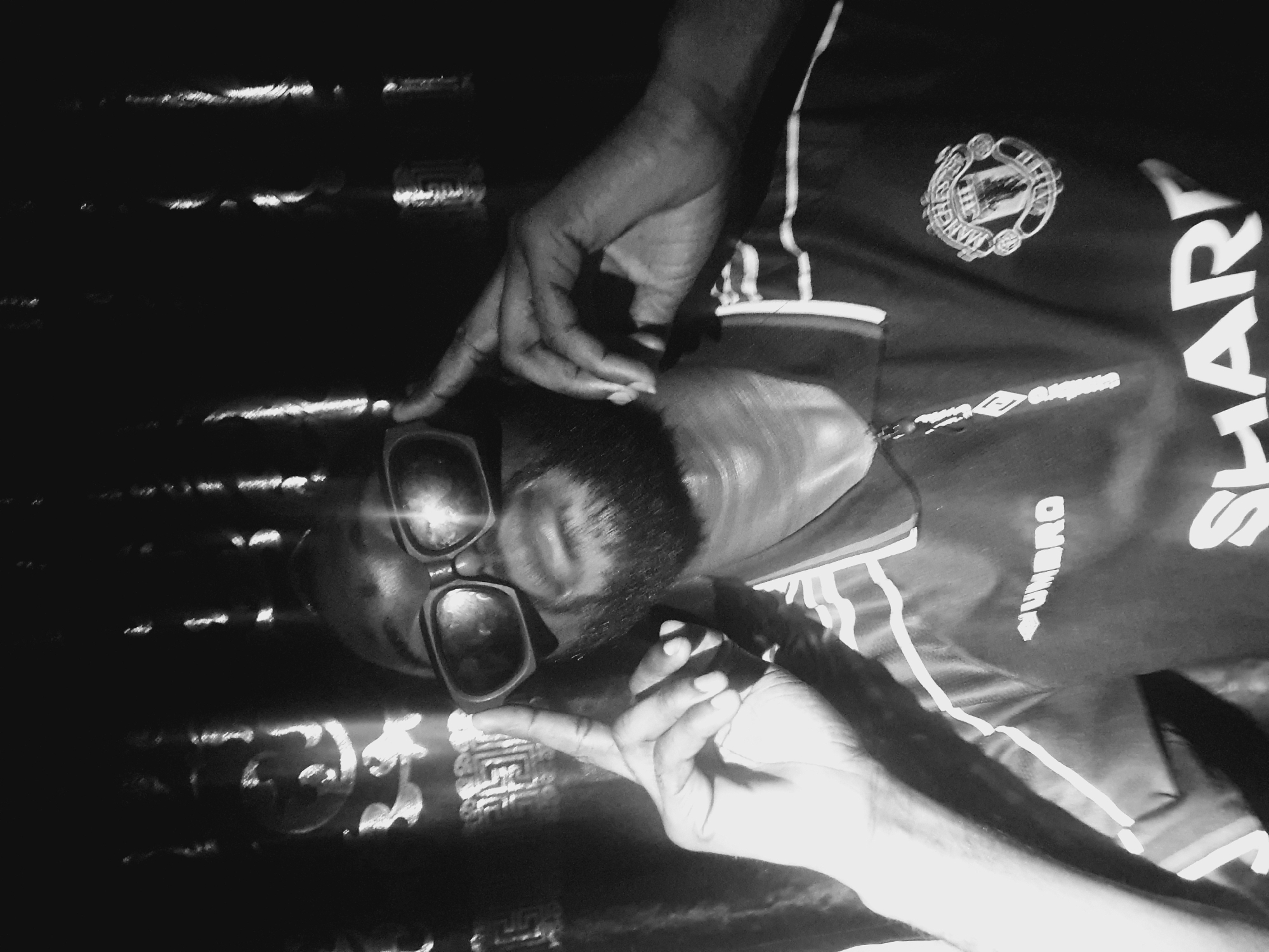The image appears to be a black and white photograph of a person wearing sunglasses and a Manchester United soccer jersey with the sponsor "SHARP" visible. The person is gesturing with their hands. The background is dark and contains reflective surfaces, potentially liquid or polished surfaces that reflect light. The Umbro logo is partially visible on the jersey. The location is not discernible in the image, and there is no visual information available to confirm that this image was taken in Lokoja, Nigeria.