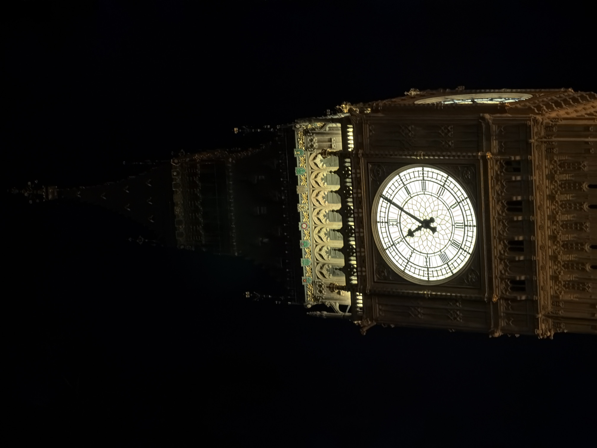 The image shows the illuminated clock face of the Elizabeth Tower, commonly known as Big Ben, in London, United Kingdom, at night. The clock is visible, indicating a time of approximately 4:52. The surrounding tower is elaborately decorated with architectural details and is illuminated with warm light. The sky is dark, providing a stark contrast to the brightly lit clock and tower. The image appears to be taken from a low angle, emphasizing the height and grandeur of the structure.