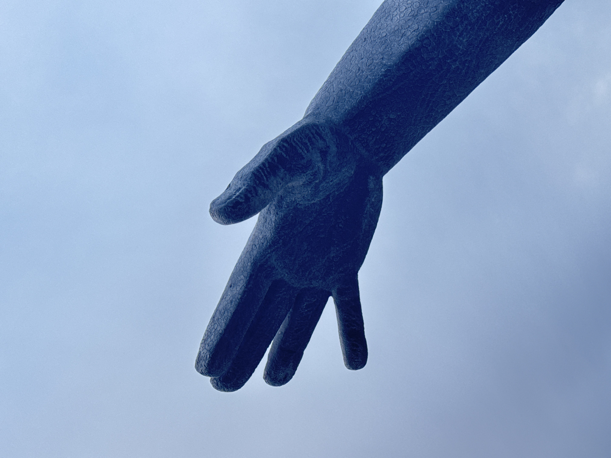 A low-angle shot depicts the hand and forearm of a bronze sculpture against a pale blue sky. The hand is positioned downwards, fingers outstretched. The texture of the bronze appears coarse, suggesting the sculpture is old or weathered. There are no discernible people or other objects in the image. It is a part of the bronze sculpture of Jesus Christ in Kazan, Russia.