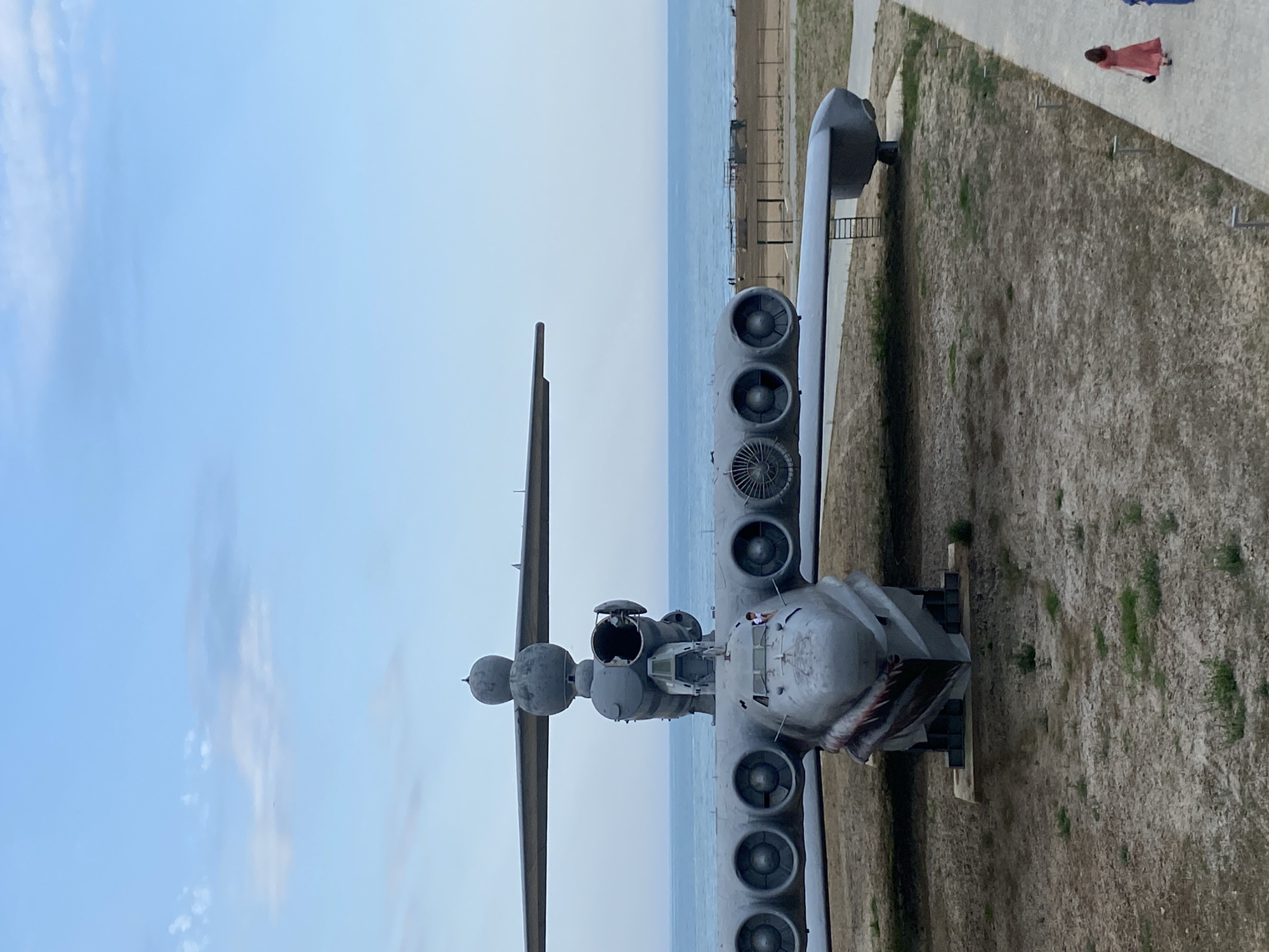 The image depicts a large, gray, winged vehicle, possibly an aircraft or a seaplane, parked on a dry, dusty ground with sparse vegetation. The vehicle has multiple engines mounted on its wings and a prominent cockpit area. In the background, a calm blue sea stretches to the horizon under a pale blue sky with scattered clouds. A sandy beach with a few structures and people is visible between the vehicle and the sea. To the right of the vehicle, a paved walkway runs alongside the scene, and two individuals, one in a red dress and another in a blue garment, are walking away from the camera on the walkway. The setting appears to be an outdoor exhibition or a museum in Arablyar, Russia, possibly a memorial site for the aircraft.