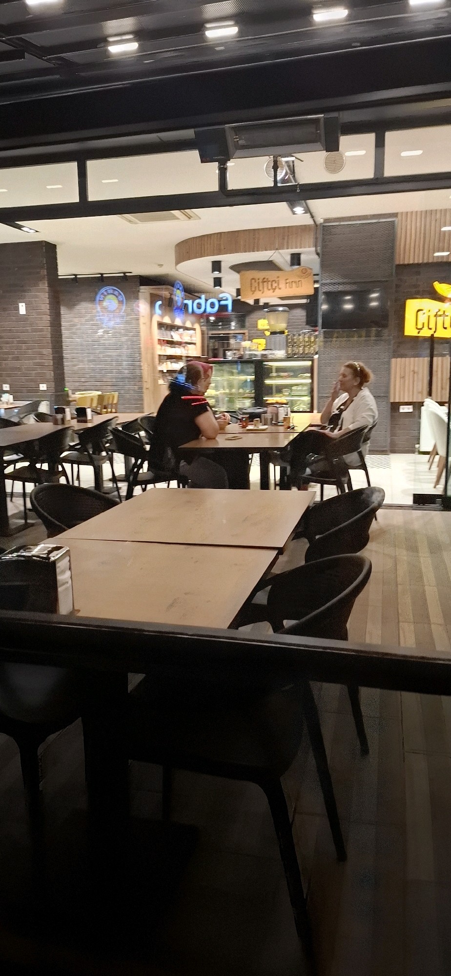 The image depicts the interior of a cafe or restaurant, possibly located in Kadıköy, Turkey, based on the "Çiftçi Fırın" signage which translates to "Farmer's Bakery" and is a common chain in Turkey. Two individuals are seated at a table in the mid-ground. The person on the left, wearing a dark shirt, is facing away from the camera, while the person on the right, in a light-colored top, is facing towards the camera and appears to be engaged in conversation or contemplation. They are seated at a wooden table with drinks and what appear to be food items. The environment is indoors, with various tables and chairs visible. The decor suggests a modern yet rustic aesthetic, with exposed brick walls and wooden accents. In the background, shelves stocked with products and display cases are visible. The lighting is artificial, creating a warm and somewhat dim atmosphere. The foreground is dominated by a dark surface, likely a table or railing, creating a sense of looking into the establishment from outside.