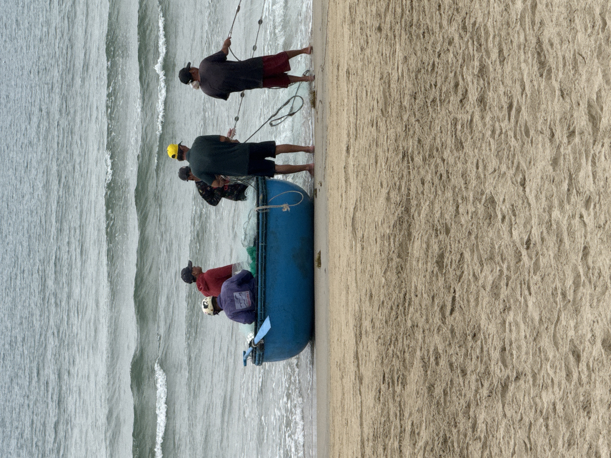 Five adult males are engaged in fishing activities on a beach in Đà Nẵng, Vietnam. A blue, round-bottomed traditional Vietnamese fishing boat (thúng chai) is partially beached at the waterline. One male, wearing a white and black patterned helmet and a purple long-sleeved shirt with a white graphic, is seated within the boat, which contains a light green fishing net. Another male, wearing a red long-sleeved shirt and a dark baseball cap, stands in the shallow water beside the boat. Three additional males are standing further in the shallow ocean water, actively pulling a large fishing net towards the shore. The central figure wears a dark green short-sleeved shirt and a bright yellow baseball cap. The male to his right wears a dark short-sleeved shirt and red shorts with a dark baseball cap. The male to his left wears a dark patterned long-sleeved shirt and a dark baseball cap. A light blue paddle is visible inside the boat. The foreground consists of light brown sandy beach, while the background features a gray-green ocean with moderate waves under an overcast sky.