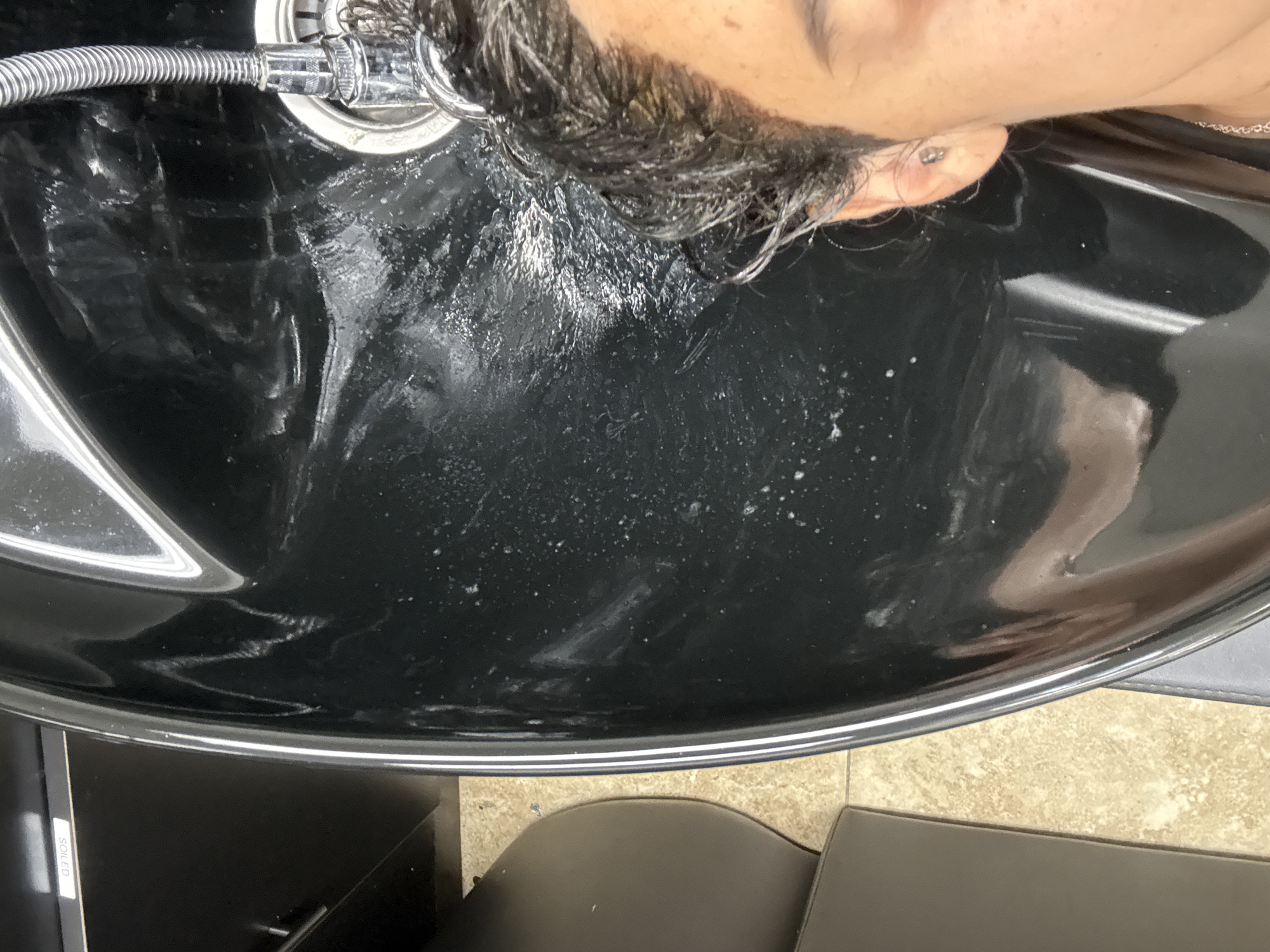 The image provides an overhead perspective of a person's head tilted back over a black, glossy salon sink, likely within a hair salon in Bellflower, United States of America. The central subjects are the person and the salon sink with its fixtures.
The person's head is positioned for a hair wash or rinse, with dark, wet hair visible, including some gray strands. A flexible, metallic shower hose is directed towards the crown of the head, and a chrome drain is present in the sink. Part of the person's light-toned forehead, cheek, and an ear with a small earring are visible.
The black sink is wet with water droplets, suds, and some residue, indicating active use. Its reflective surface shows blurred reflections of what appears to be overhead lighting or other salon equipment. Below the sink, parts of a dark chair or stool are visible, alongside a beige tiled floor. A label on a dark surface to the bottom left reads "SOILED" vertically. The scene captures the process of hair washing in a professional setting under indoor, artificial lighting.