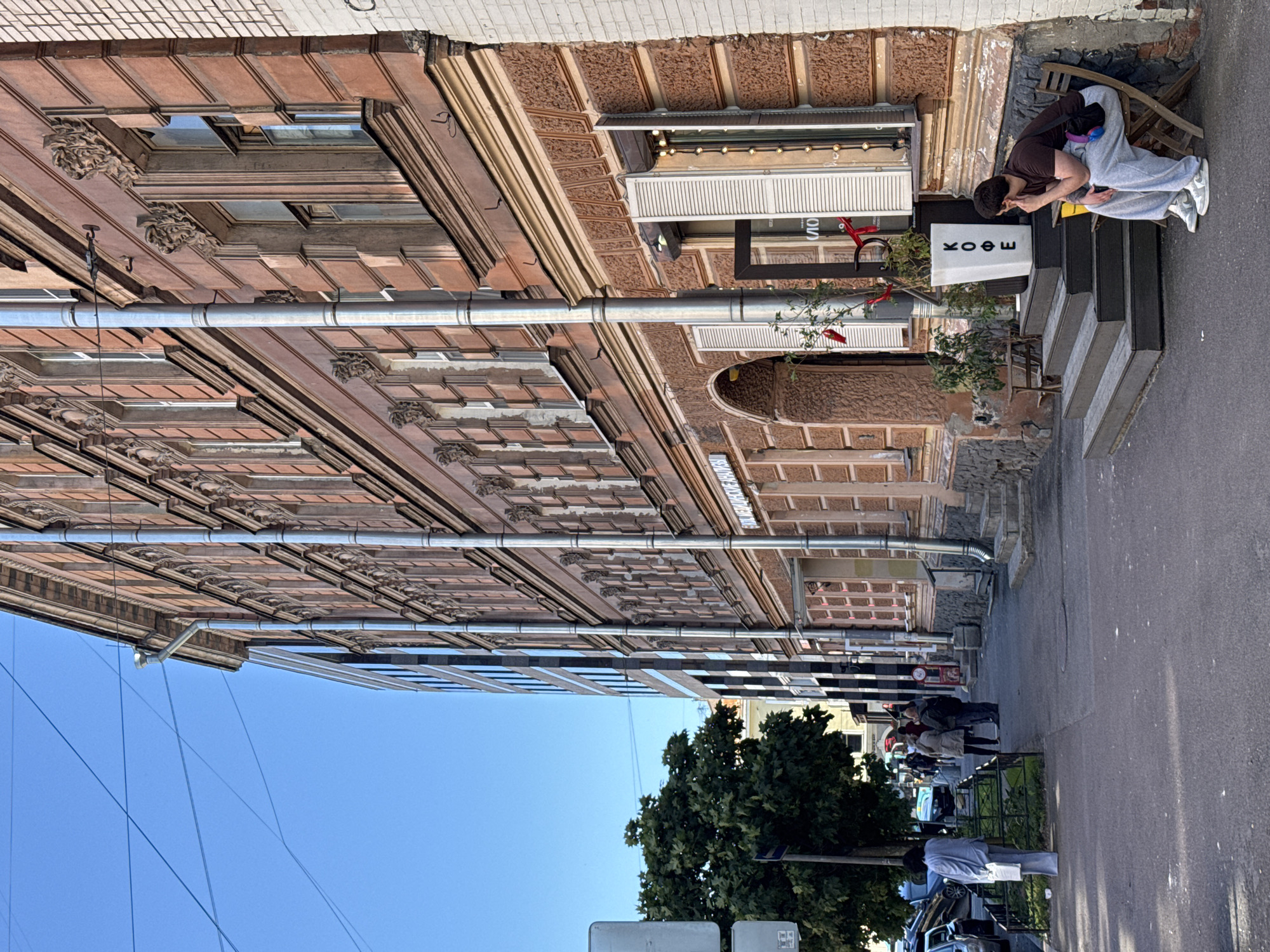 A full, eye-level shot captures a street scene in an urban environment under a clear blue sky. On the right, a weathered, reddish-brown building with ornate architectural details stands prominently. The building features multiple windows with white shutters, some of which are open, revealing interior lights. Downspouts run vertically along the facade. In front of the building, a set of wooden steps leads up to an entrance. A white planter box with the Cyrillic word "КОФЕ" (coffee) in black lettering sits at the top of the steps. To the left of the building, a person sits on a wooden chair, leaning forward with their hand on their chin, appearing contemplative.
The sidewalk in front of the building is paved, and further to the left, a tree with dense green foliage casts a shadow. A metal fence lines a grassy area next to the sidewalk. Beyond that, a street is visible with several parked cars and pedestrians walking. Another building, modern and with a glass facade, stands to the left of the older building, creating a contrast in architectural styles. Overhead, power lines crisscross the sky. The overall impression is of a quiet street on a sunny day.