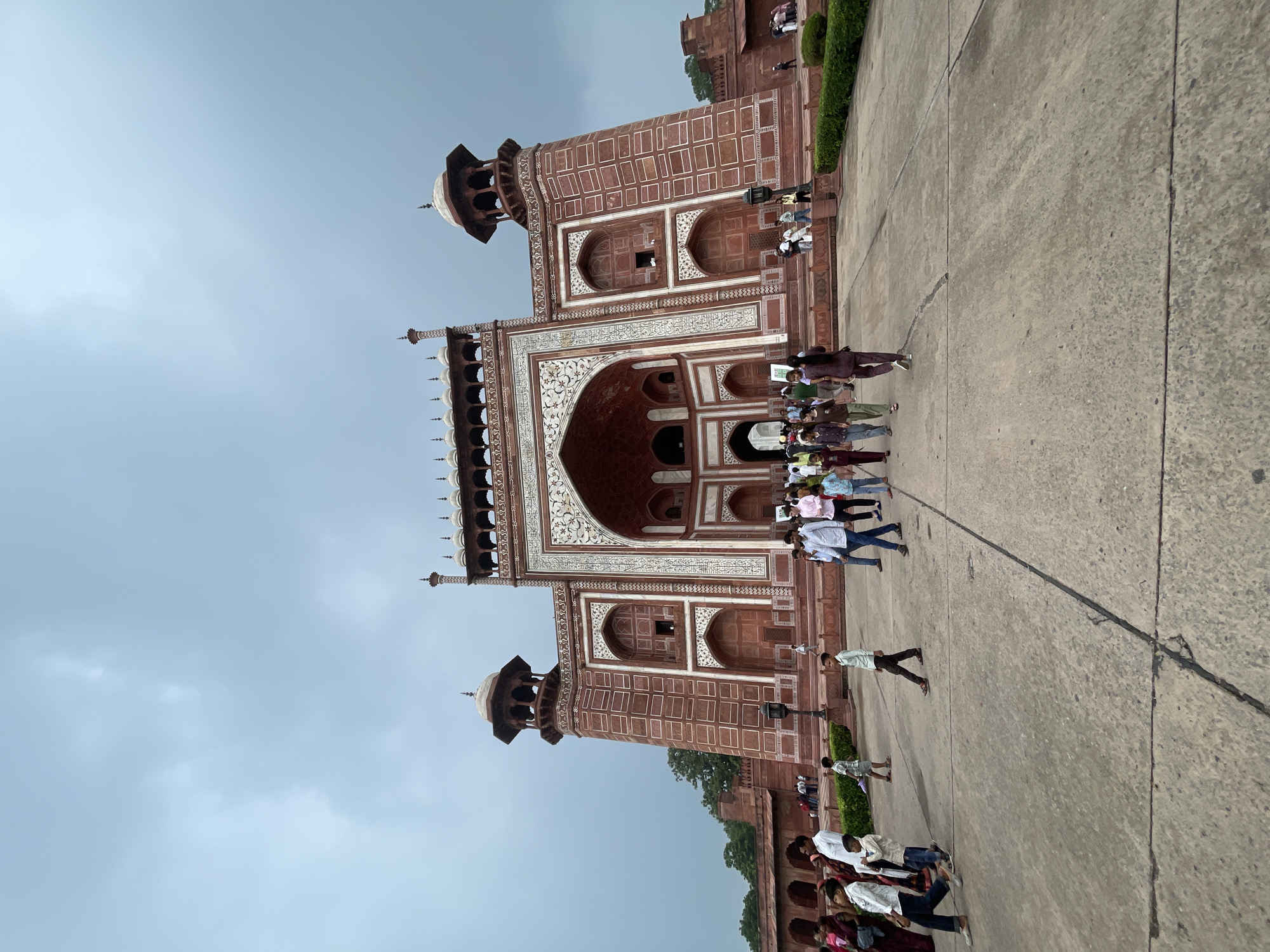 This is a wide-angle, low-angle, eye-level shot of the Taj Mahal's South Gate in Agra, India. The gate is a large, red sandstone structure with intricate white marble inlay work. It features a central archway, flanked by smaller arched doorways and ornamental minarets. A crowd of people, dressed in casual, colorful clothing, are walking towards and away from the gate. The sky above is overcast with dark, dramatic clouds. The foreground is a large, paved courtyard, and a trimmed green hedge is visible to the right. The overall atmosphere is one of tourism and historical grandeur.