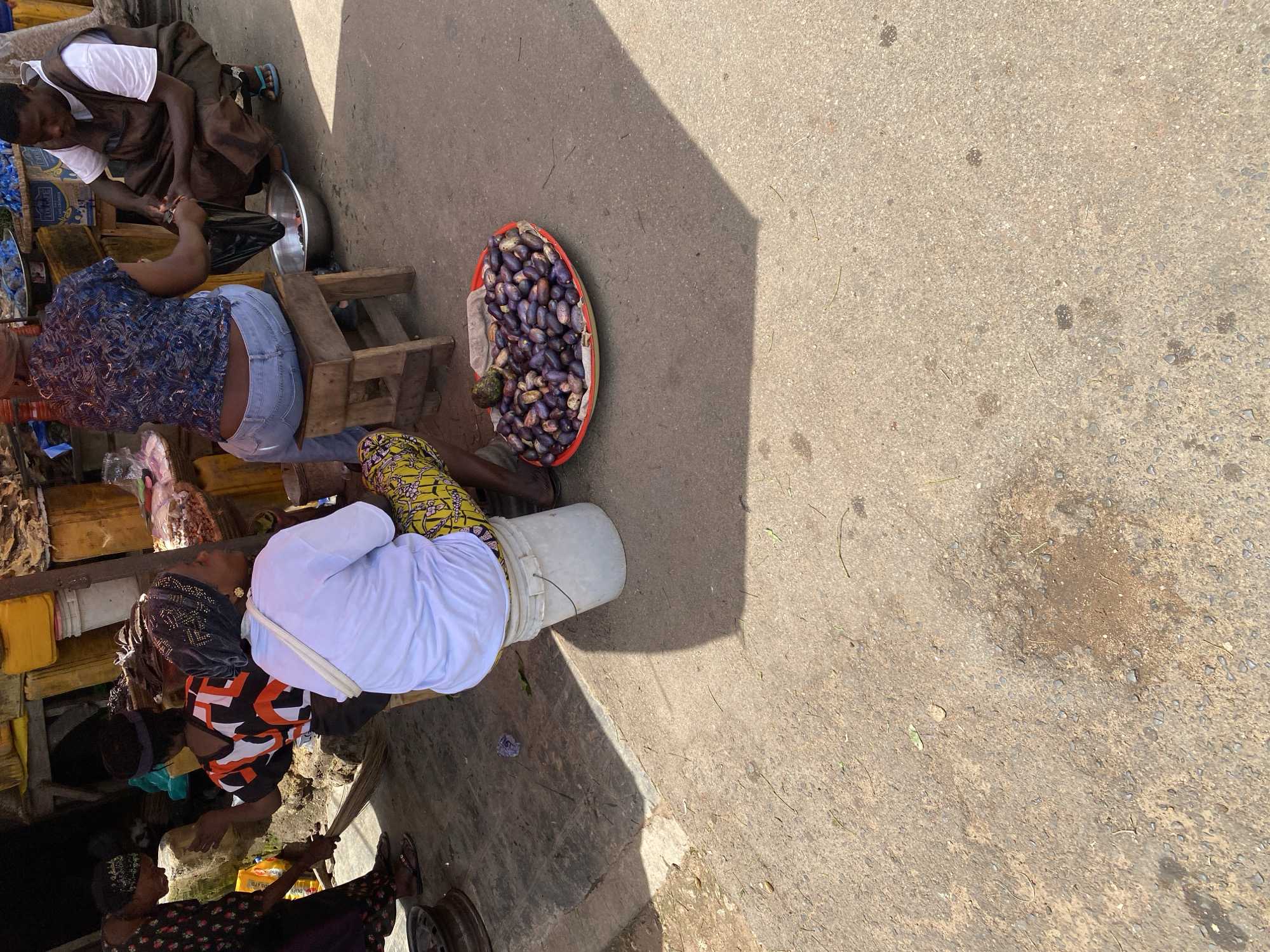 The image depicts a street market scene in Jos, Nigeria. In the foreground, a person wearing a white shirt and yellow patterned pants sits on a white bucket. To the right of this individual is a red plastic bowl filled with numerous purple fruits. In the background, several other people are visible. One person is sweeping the ground with a broom, while another is interacting with a man who is crouched down. The environment appears to be an outdoor market with various stalls and goods, suggesting a bustling commercial area. The ground is paved with rough asphalt, and shadows from overhead structures are cast across the scene.