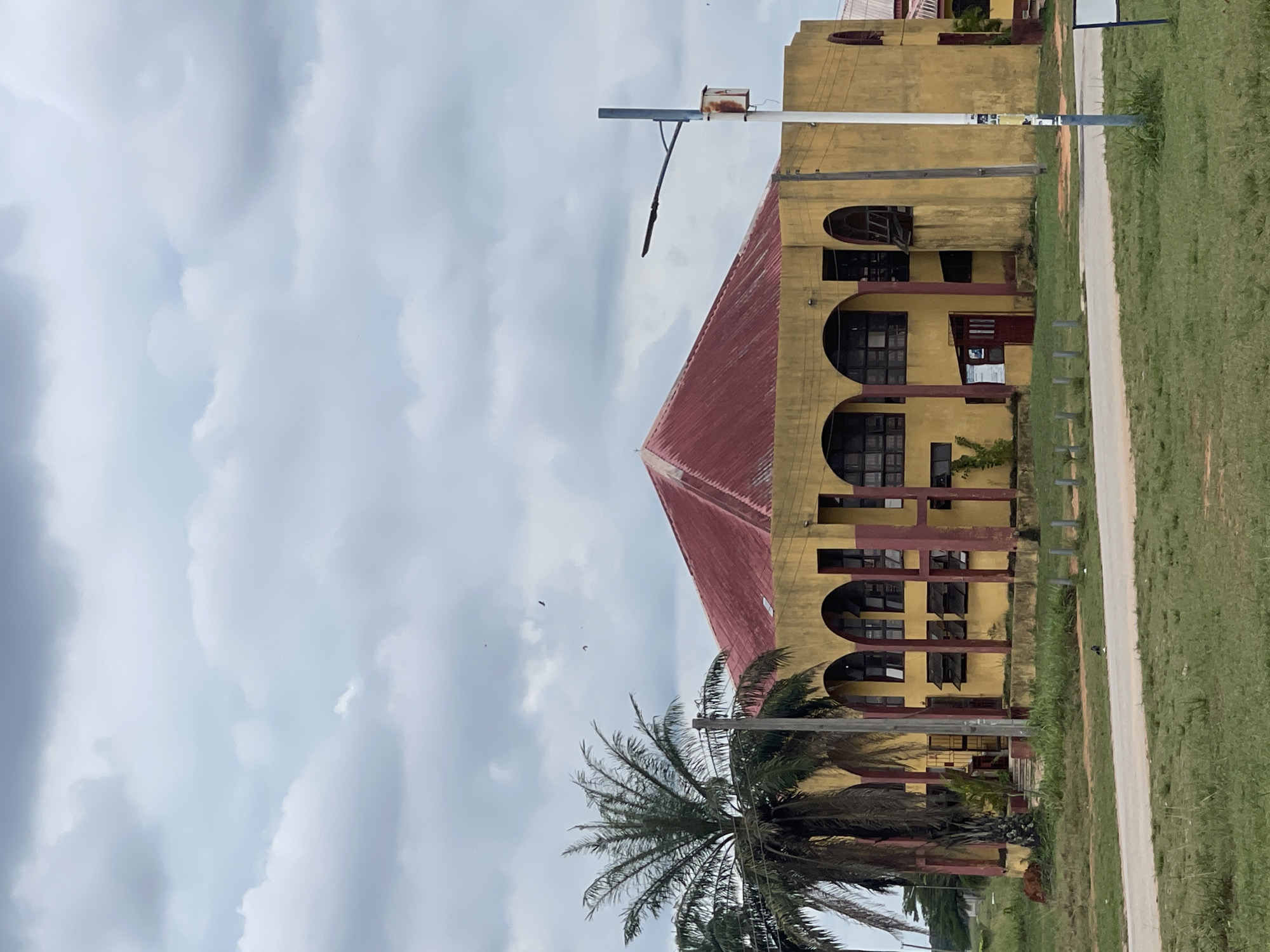 The image shows a yellow building with a red roof, featuring arched windows and columns, likely located in Abraka, Nigeria. A palm tree stands in front of the building. A road and grassy area are to the right. Overhead, a cloudy sky is visible.