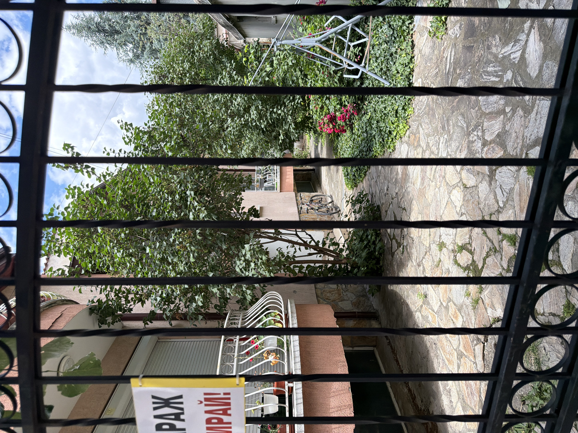 The image captures a courtyard scene viewed through a black metal fence. The courtyard is paved with irregular flagstones and is bordered by lush greenery, including bushes and trees. A pink-colored building with a balcony is visible on the left, featuring white railing and potted plants. A yellow sign with Cyrillic text is attached to the fence, partially obscuring the building. Two bicycles are parked near the building. To the right, a light blue swing set is situated amidst dense green ground cover and flowering pink bushes. The sky above is partly cloudy. This scene is located in Pazardzhik, Bulgaria, likely a residential area given the presence of the building and courtyard features. There are no people visible in the image.