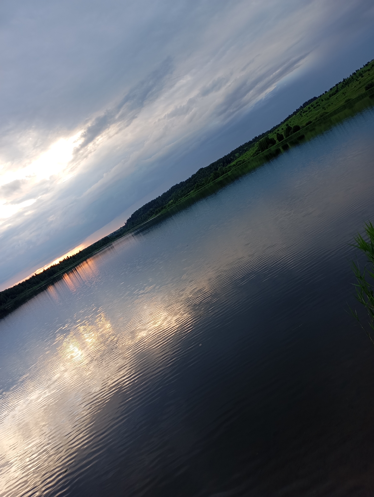 The image depicts a body of water, likely a lake or river, with a shoreline covered in green vegetation and trees. The sky above is filled with dramatic clouds, with a bright patch of sunlight breaking through on the left side. The water reflects the sky and the distant shoreline. The scene is captured at an angle, with the shoreline and sky running diagonally across the frame. The overall impression is of a natural landscape at dusk or during a cloudy day. The location is identified as Ryabki, Russia. There are no people or discernible human activity visible in the image. The water is calm, with gentle ripples on its surface.