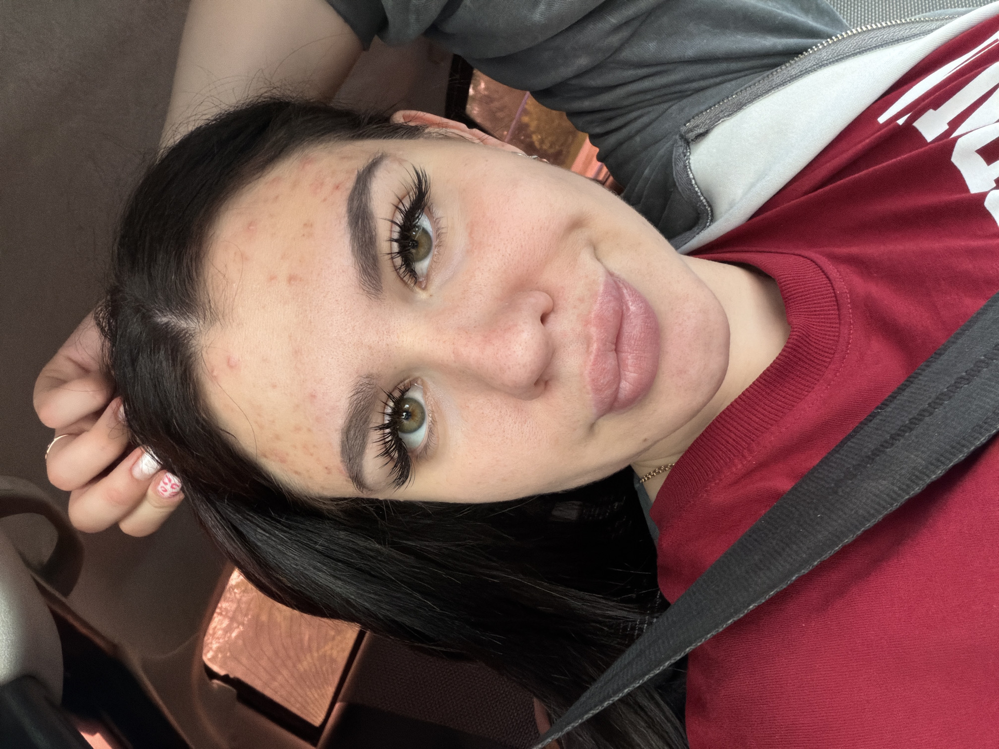 The image shows a close-up, frontal portrait of a young woman with dark hair, green eyes, and full lips. She is wearing a red t-shirt and a grey zip-up hoodie. A black seatbelt is visible across her chest. Her hand is resting on her head. There are several blemishes on her forehead and cheeks. She is indoors, possibly in a vehicle, as evidenced by the window tinting and what appears to be part of a car interior in the background. The image does not contain any information that could place the location in Chaikovsky, Russia.
