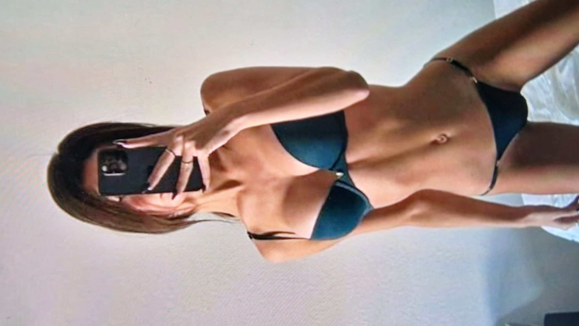 A young woman is captured in a selfie, holding a dark-colored smartphone in her right hand to obscure her face. She is wearing a dark-colored bra and a matching thong, with her left arm extended. The background is a plain, light-colored wall. The setting appears to be indoors, possibly a bedroom, with a white surface visible in the lower portion of the frame, suggesting a bed. There is no indication of the city of Aksu, China, or any urban environment in the image.