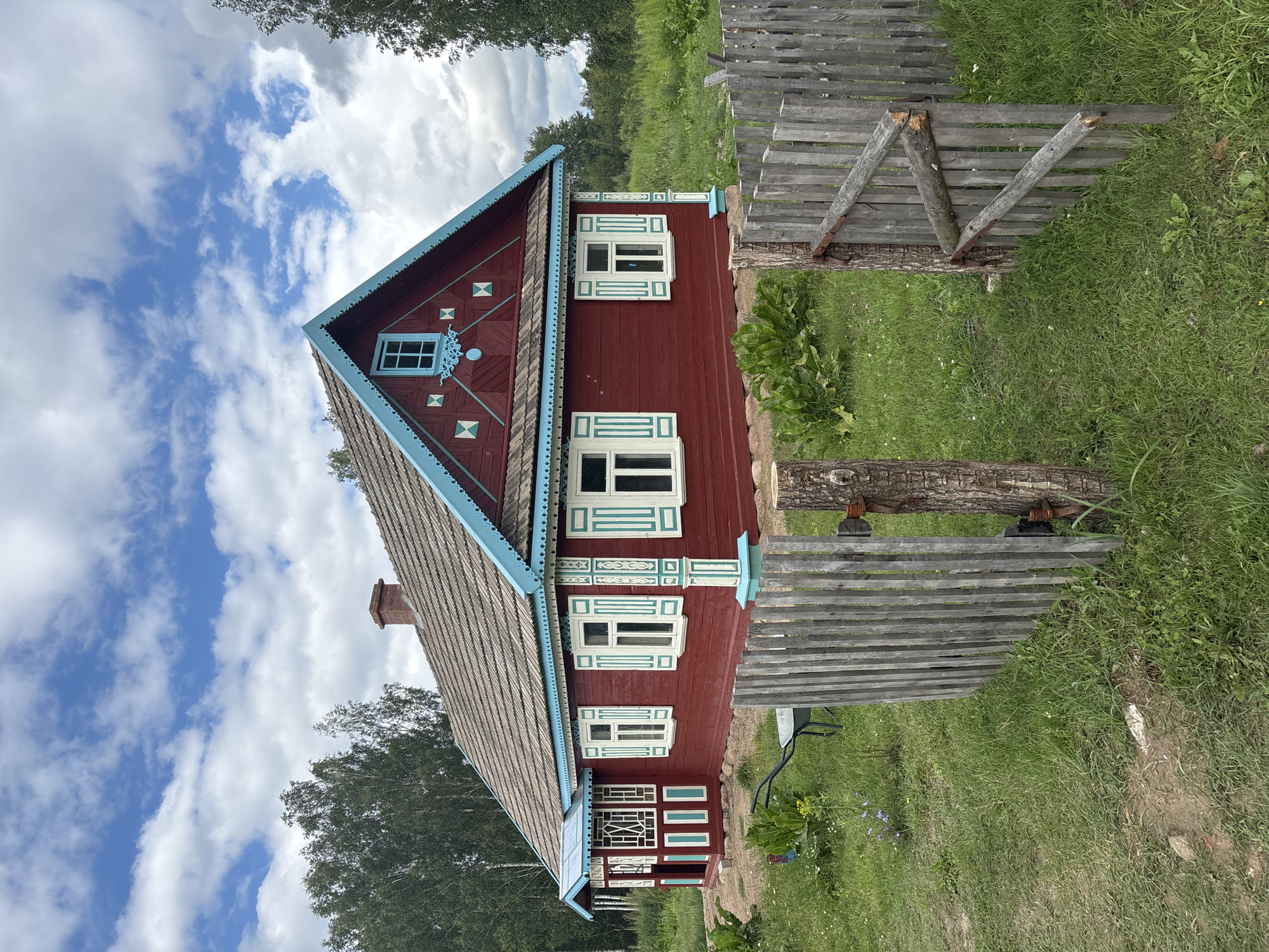 This media file displays a traditional, brightly painted red wooden house with a blue-trimmed roof and decorative window frames, set in a lush, green, grassy landscape. The house features several windows with white shutters and intricate blue and white trim. A weathered wooden fence runs along the side of the property, and a chimney is visible on the roof. The sky is a vibrant blue with scattered white clouds, suggesting a pleasant, possibly sunny day. Trees can be seen in the background, indicating a rural or semi-rural setting. There are no people or animals visible in the image, and no text is present. The scene evokes a sense of rustic charm and tranquility. The orientation of the image is portrait and rotated 90 degrees clockwise.