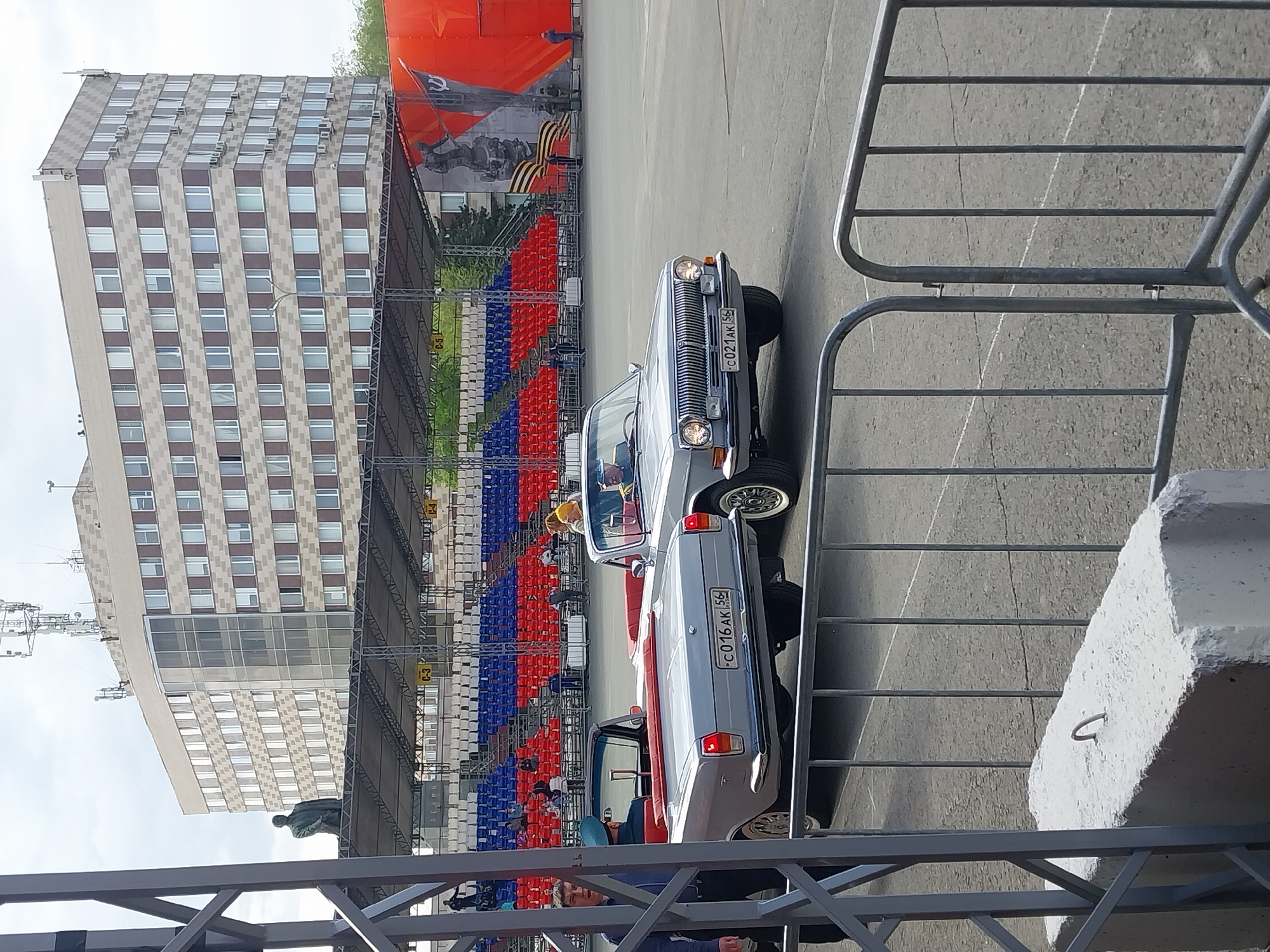 The image captures a scene at a public event in Orenburg, Russia. In the foreground, a metal fence obscures a portion of the view. Behind the fence are two vintage silver cars, possibly participating in a parade or display. A spectator is visible in one of the cars. Red, white, and blue seating lines a portion of the parking lot. In the background, a large Soviet-era mural is visible and the buildings of the city of Orenburg.