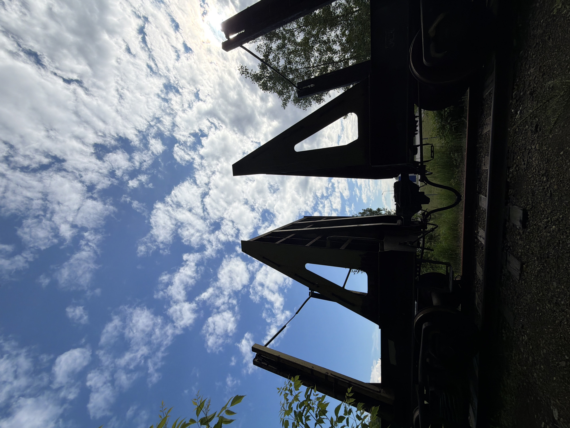 The image captures an upward-looking perspective of heavy industrial or railway equipment against a bright, partly cloudy sky during the daytime. The scene is set in Chaikovsky, Russia.
The upper two-thirds of the photograph are dominated by a vibrant blue sky streaked with numerous white, fluffy clouds. The sun is visible in the upper right quadrant, casting a strong glare that diffuses through the clouds.
In the foreground, two prominent, dark, triangular structures rise sharply into the frame, silhouetted against the bright sky. These appear to be hinged panels or ramps, possibly belonging to an open railway wagon or a specialized flatcar. Their angular forms create stark geometric patterns against the natural backdrop of the sky. Bits of green foliage, including leaves and branches, are visible around these structures, some also silhouetted, suggesting vegetation growing in and around the equipment.
The lower portion of the image reveals the darker, less distinct details of the industrial structure. Elements of a train's undercarriage, including a large wheel, are discernible in deep shadow. To the right, a dark, gravelly surface, likely a railway bed, is partially visible. The overall impression is one of a functional or formerly functional industrial site, possibly a railway yard, where nature has begun to reclaim its space. No specific activity is depicted, and no discernible text is visible.