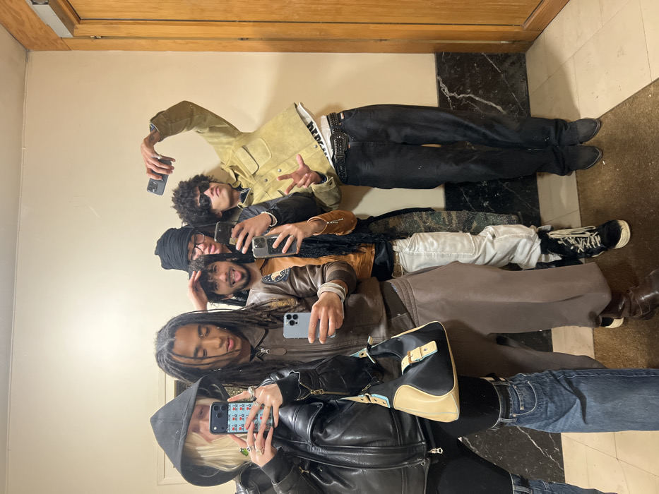 The scene depicts five individuals posing for a photograph within an indoor hallway or vestibule in Paris, France. The environment features plain, light-colored walls above dark marble wainscoting and a light-toned floor with a brown mat in the foreground. A wooden door and doorframe are visible on the right.
From left to right:
1. A blonde individual, wearing a black hooded jacket and black pants, holds an iPhone in a green-patterned case aloft, appearing to capture the image. A black and beige shoulder bag with light blue accents is slung over their left arm.
2. A male with dreadlocks, dressed in a brown zippered jacket and loose-fitting brown pants, holds a light-colored iPhone at waist level.
3. A male with dark, curly hair and a beard, wearing a brown jacket over a dark top with a patterned scarf, extends his tongue slightly. He holds an iPhone at chest level and places his left hand on the head of the individual next to him. He wears white pants and black boots.
4. A male in a black headwrap and eyeglasses, wearing a dark jacket over a white shirt, is partially obscured by the individual to his left. He also holds a smartphone. His pants feature a camouflage or patterned design.
5. A male with dark, curly hair and sunglasses, wearing a tan suede-like jacket over a white t-shirt, holds an iPhone high above his head with his right hand, extending his left hand in a "peace" sign. He wears a prominent belt buckle with dark pants and black pointed shoes.
All individuals are looking generally towards the point where the image is being captured, suggesting a mirror selfie or a group pose for another camera not visible in the frame.