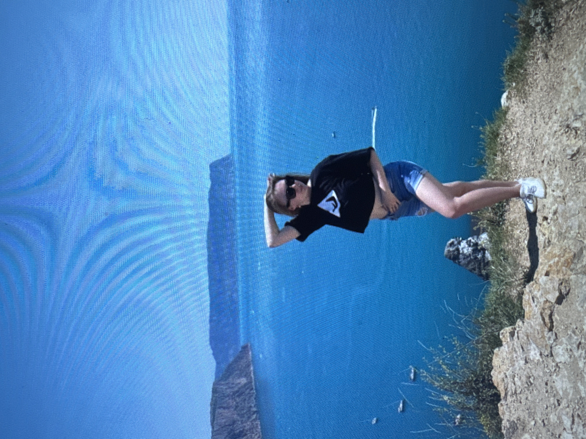 A young woman with blonde hair stands on a cliff overlooking a bright blue body of water. She is wearing a black crop top with a white triangular logo, denim shorts, and white sneakers. Her right arm is raised, with her hand resting on her head. She is wearing sunglasses. The water is calm and dotted with several small boats. In the background, distant mountains rise from the sea under a clear blue sky. The cliff face is rocky and covered with sparse vegetation. The image appears to be taken on a sunny day.
