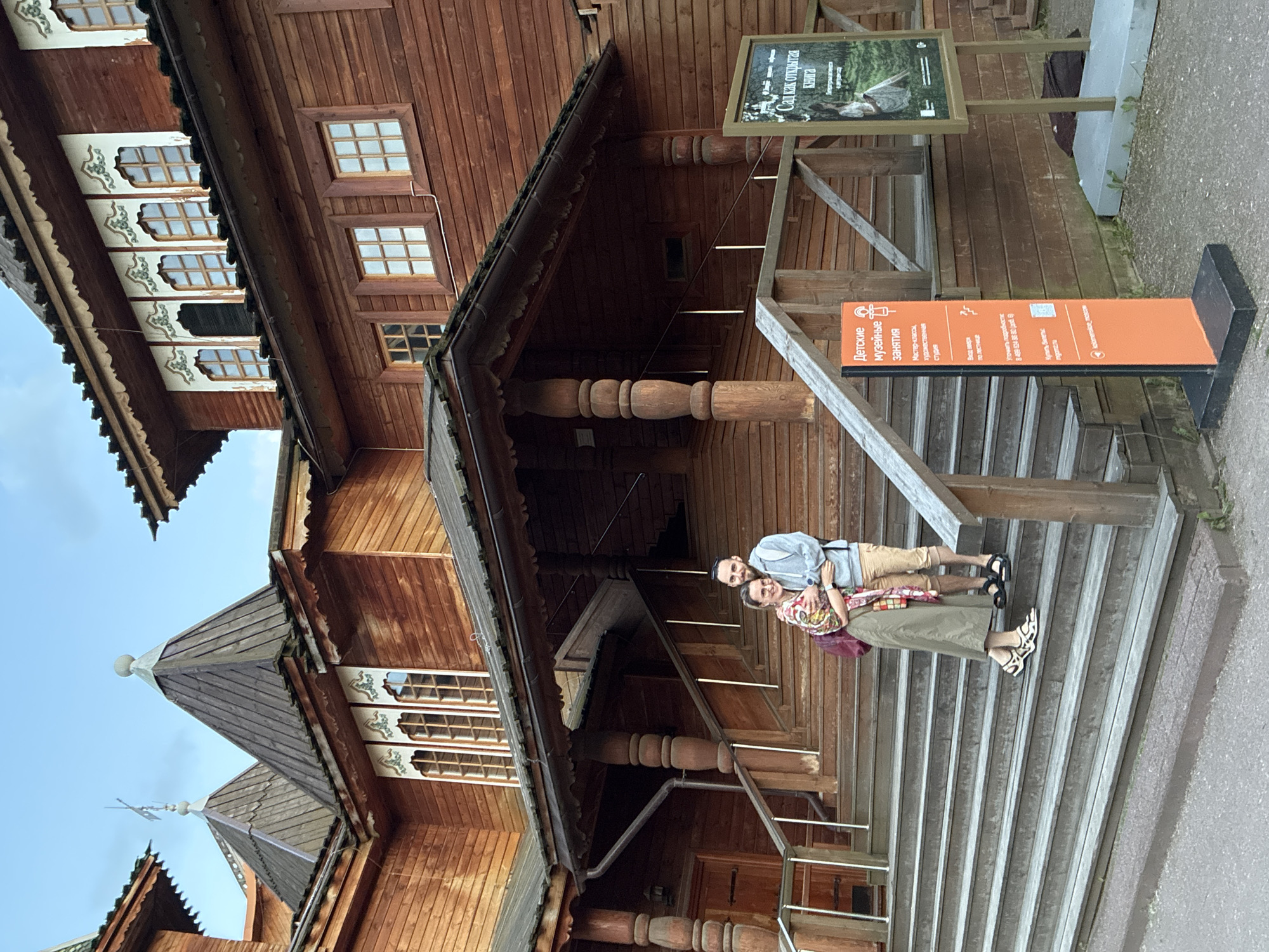 A photograph depicts two smiling individuals, a man and a woman, posing on wooden stairs in front of a grand, multi-story wooden building. The architecture features traditional Russian styling with reddish-brown timber construction, numerous windows with grid panes, and ornate decorative elements under the eaves. Pointed, spire-like roof structures are visible towards the top left, characteristic of historical Russian buildings.
The man, positioned on the left, wears a light blue button-down shirt, khaki shorts, and dark sandals, sporting a beard and a happy expression. His arm is around the woman, who stands to his right. She is dressed in a colorful patterned top, a long olive-green skirt, and distinctively patterned sandals. Both are looking directly at the camera, suggesting they are visitors enjoying their outing.
To the right of the stairs, an orange vertical sign displays information in Russian, translating to "Children's museum activities," "Master classes, art studio," and "Entrance up the stairs." It also lists a phone number for inquiries and information about buying tickets. Above it, a framed sign with a nature image bears the Russian text "Сад как открытая книга" (Garden as an open book).
The scene is set outdoors during what appears to be daytime, under a light blue sky, possibly with some overcast conditions given the soft lighting. The overall impression is one of a pleasant visit to a cultural or historical site, likely a museum or garden, consistent with the Moscow, Russia context.