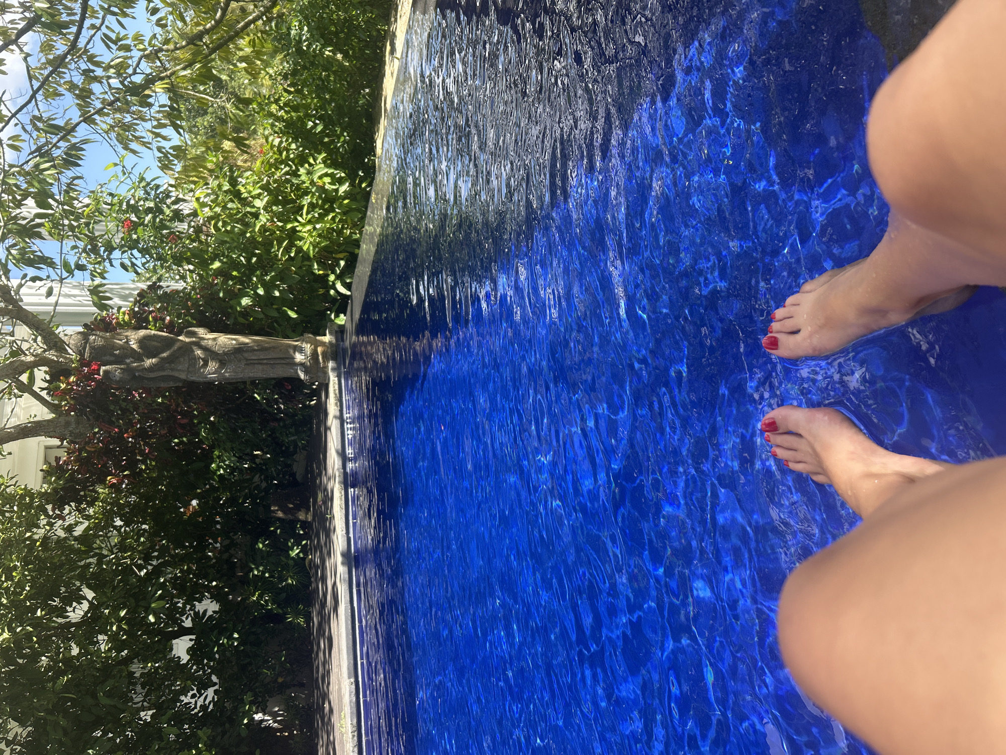 A person's legs and feet with red nail polish are visible on the edge of a swimming pool filled with blue water. The pool's edge is constructed with dark-colored tiles. The background includes green foliage with glimpses of a white structure and what appears to be a stone statue or sculpture. It is not possible to confirm the location in the city of Kediri, Indonesia, based on the visual information provided.